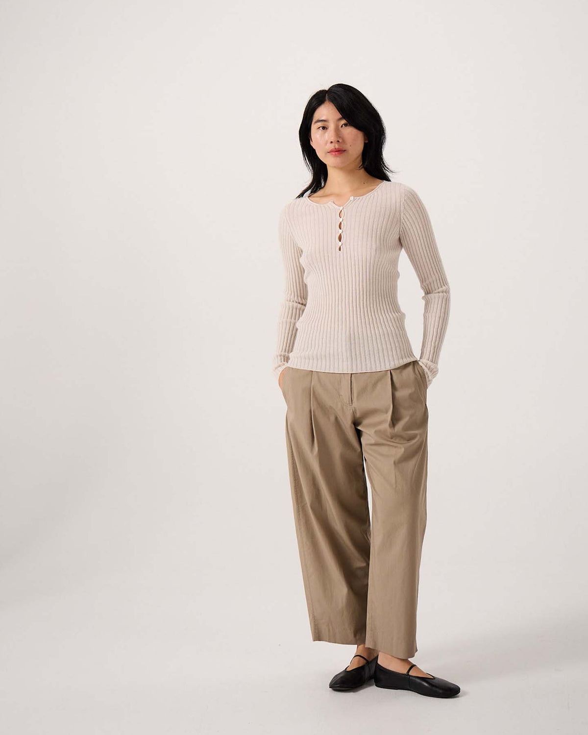 A petite woman stands against a plain background wearing the Form Top – Ivory with loose tan trousers and black flats, her hands in her pockets.