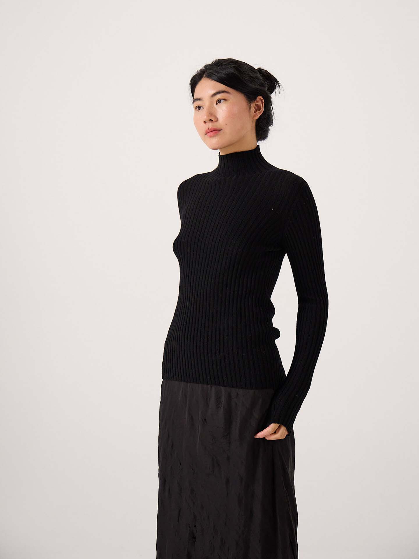 A petite person stands against a plain background, wearing the Heart Turtle Neck – Black and a black skirt, looking slightly to the side.