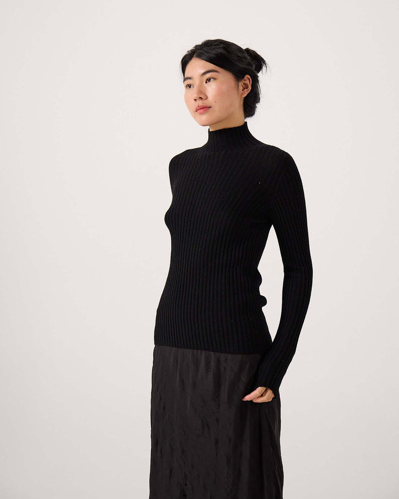 A petite person stands against a plain background, wearing the Heart Turtle Neck – Black and a black skirt, looking slightly to the side.