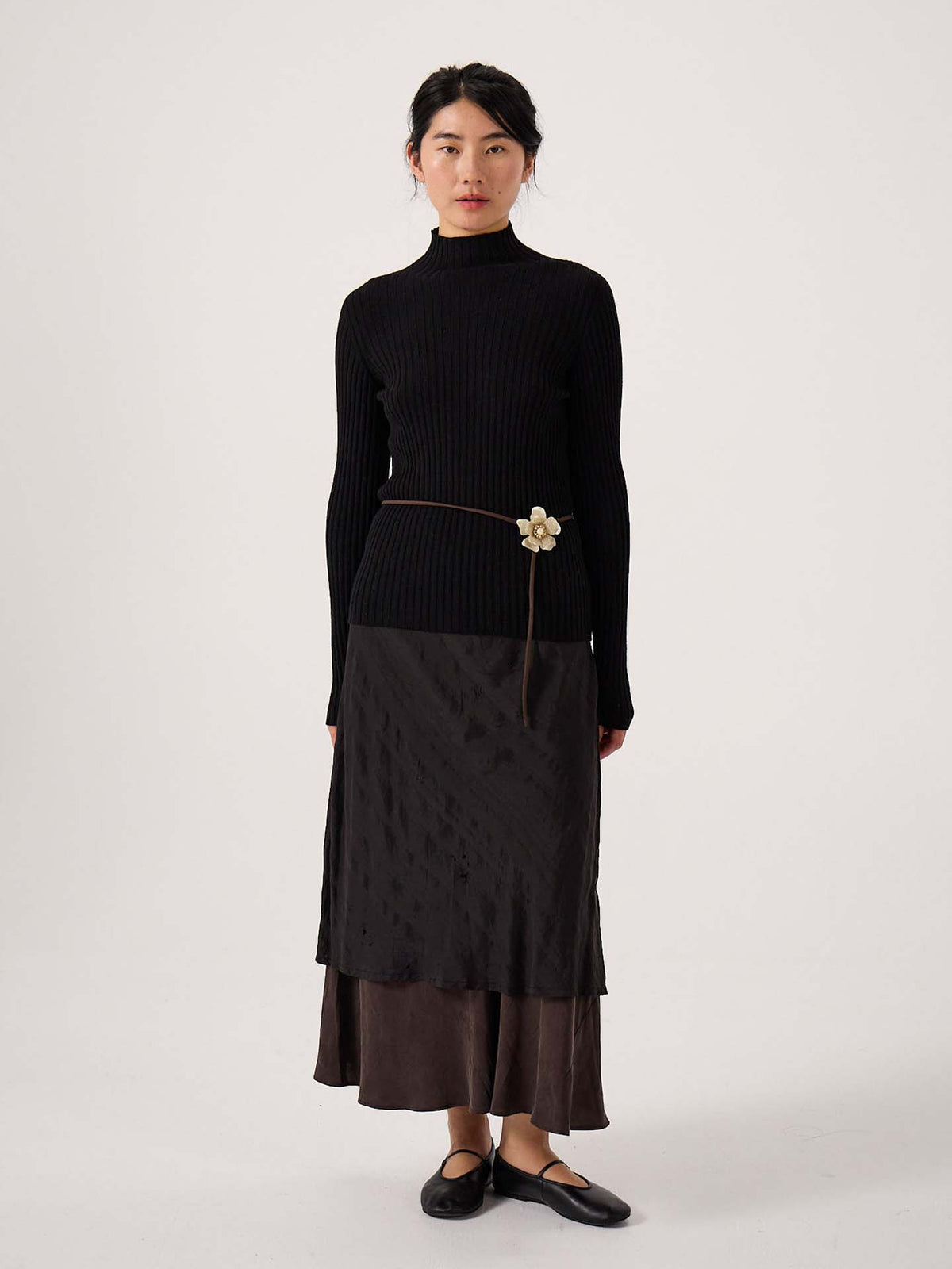 A petite woman stands facing forward wearing the Heart Turtle Neck – Black, paired with a dark layered midi skirt, black flats, and a thin belt featuring a flower detail.