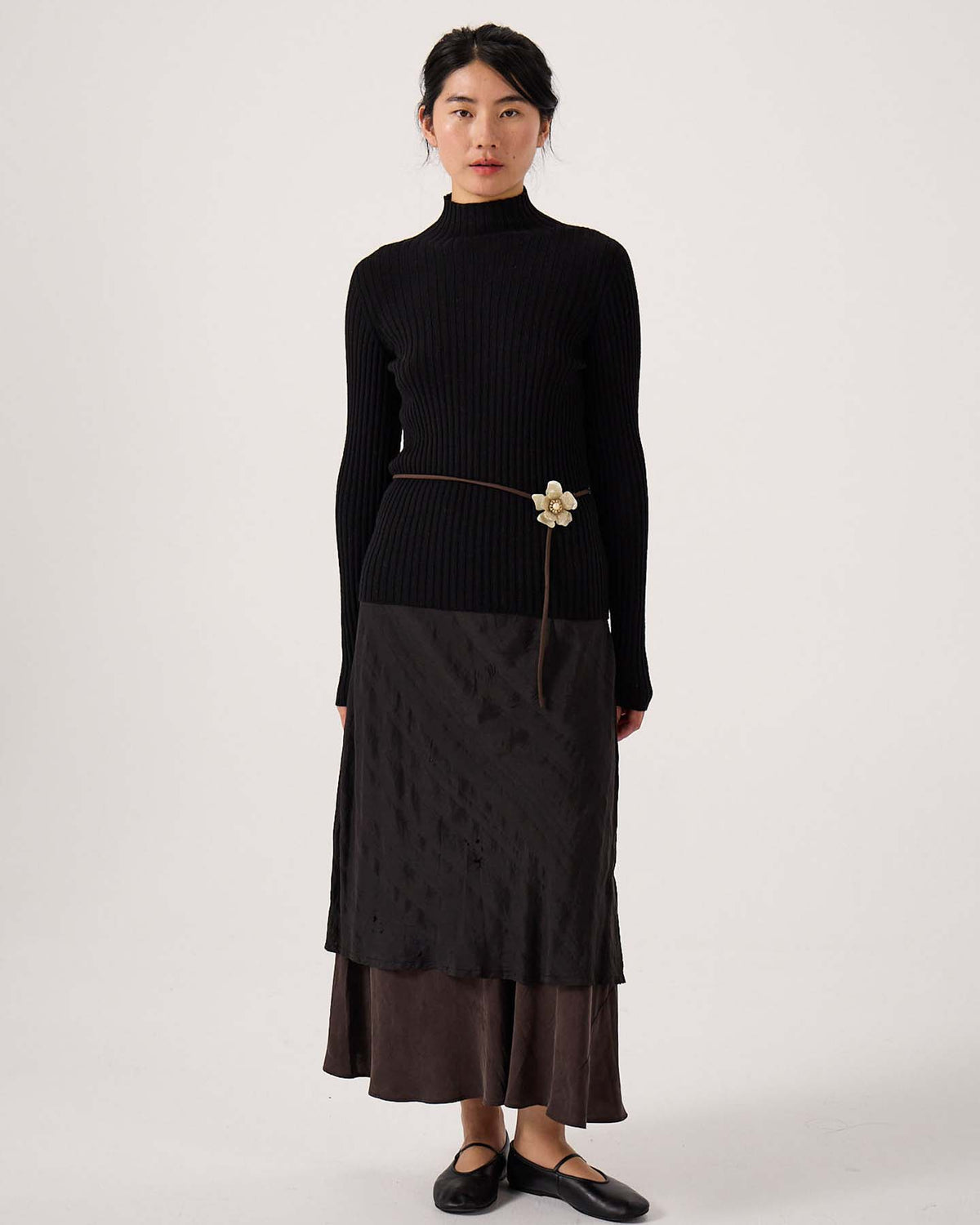 A petite woman stands facing forward wearing the Heart Turtle Neck – Black, paired with a dark layered midi skirt, black flats, and a thin belt featuring a flower detail.