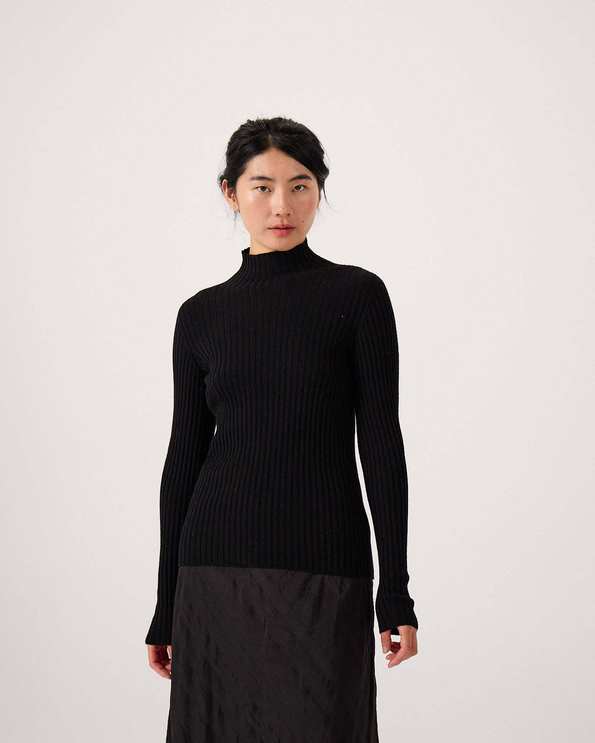 A petite woman stands against a plain background wearing the Heart Turtle Neck – Black, a stretchy, ribbed, long-sleeved turtleneck sweater paired with a matching black skirt.