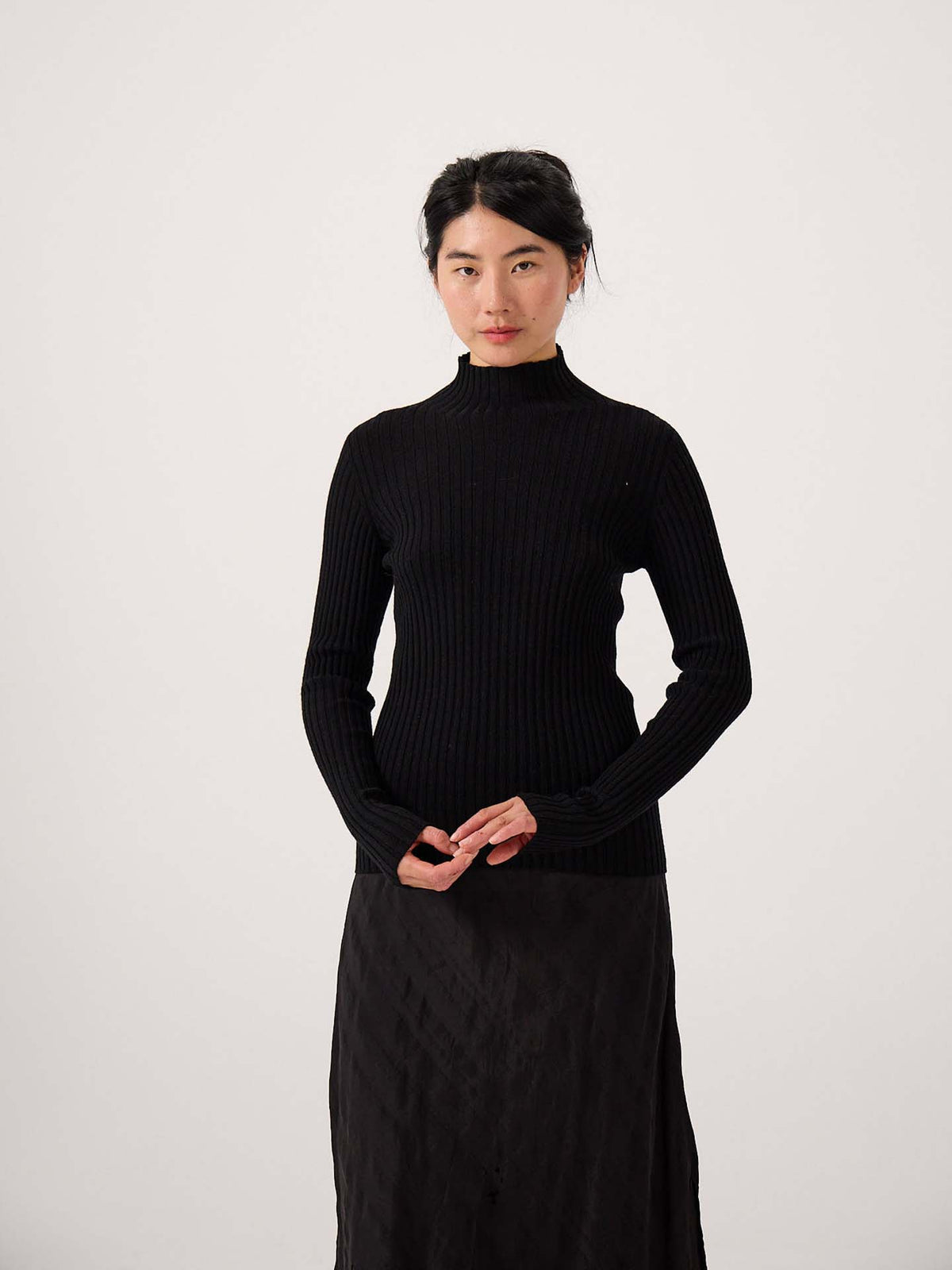 A petite woman with dark hair in a low bun wears the Heart Turtle Neck – Black and a black skirt, standing against a plain light background, highlighting the ribbed texture.