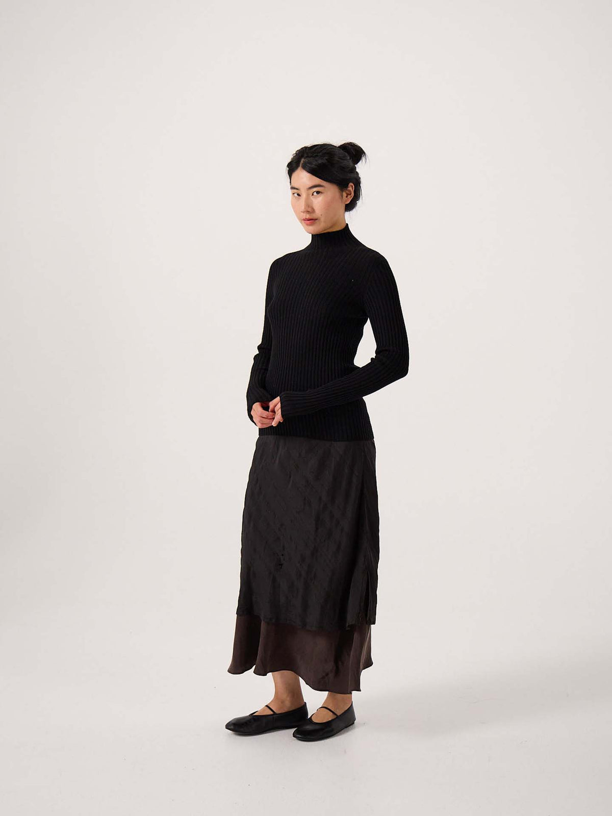 A petite person stands against a plain background, wearing the Heart Turtle Neck – Black, a layered dark skirt, and black flats, with hands lightly clasped in front.