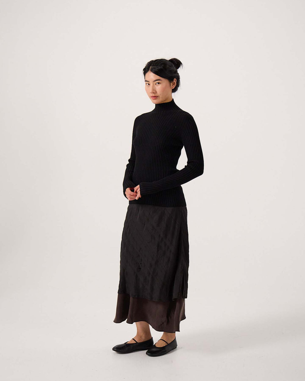 A petite person stands against a plain background, wearing the Heart Turtle Neck – Black, a layered dark skirt, and black flats, with hands lightly clasped in front.