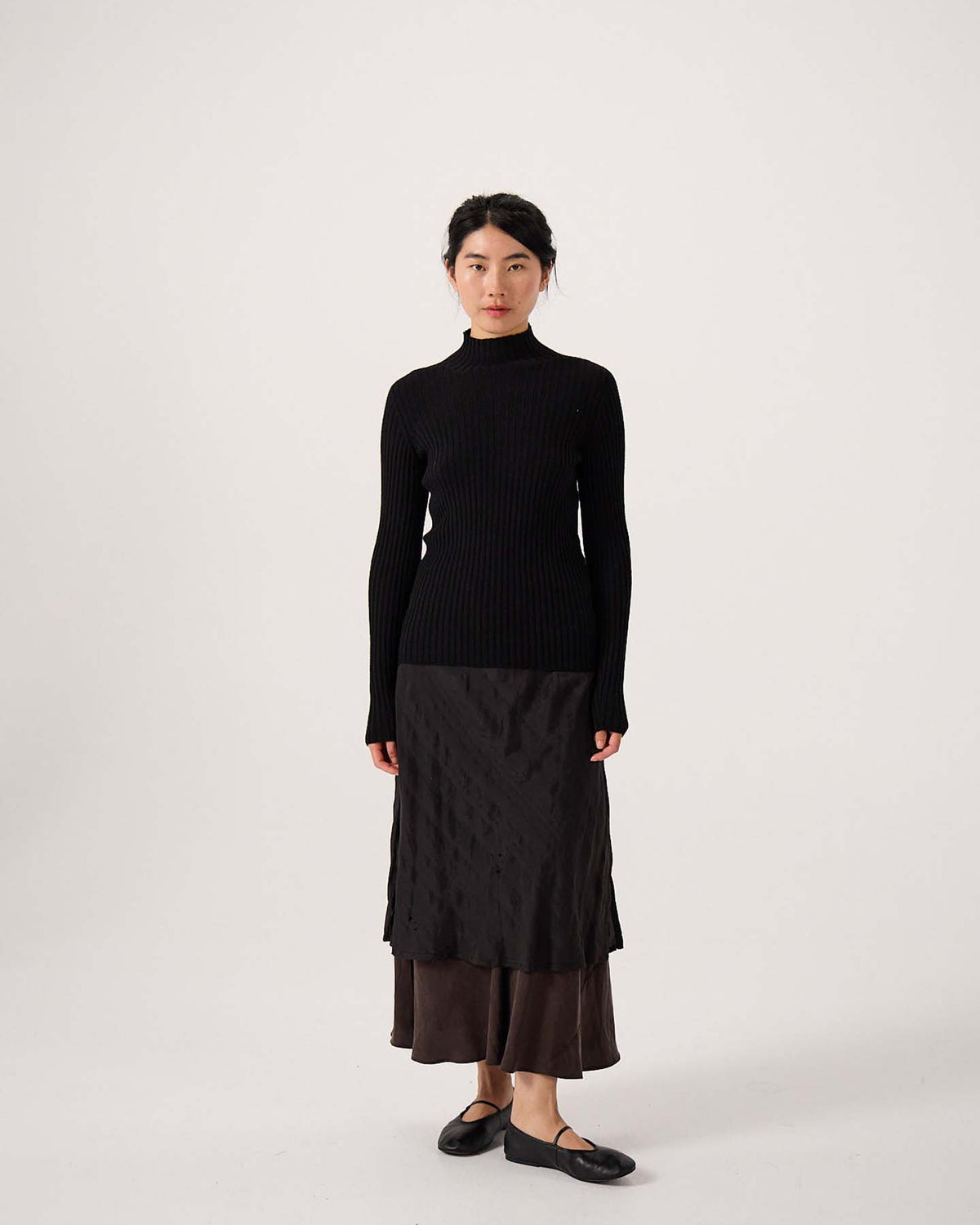 A petite woman stands facing forward against a plain light background, wearing the Heart Turtle Neck – Black sweater, a dark layered skirt, and black flats.