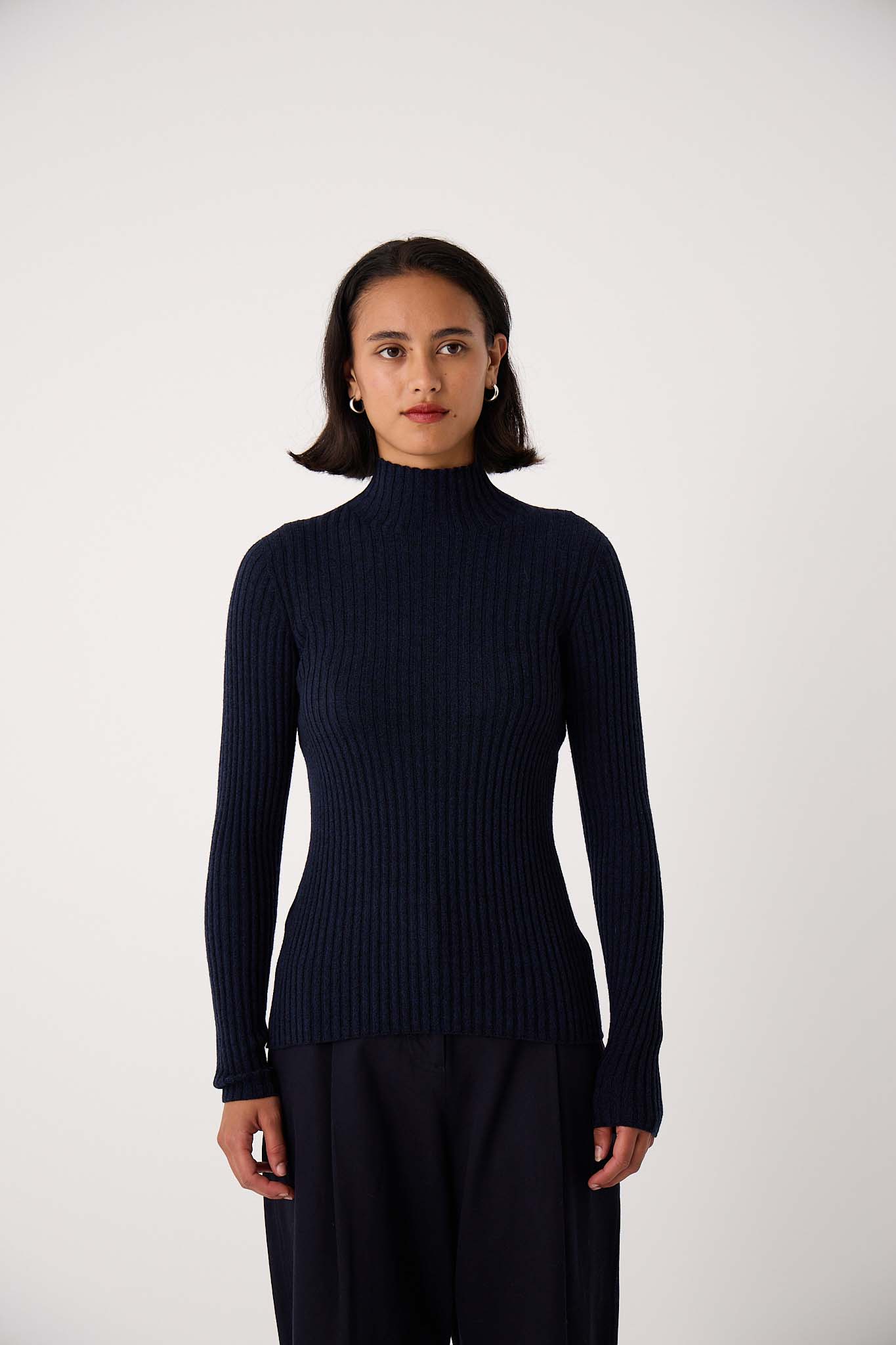 A woman faces forward against a plain light background, wearing the Heart Turtle Neck – Ink sweater with dark pants.