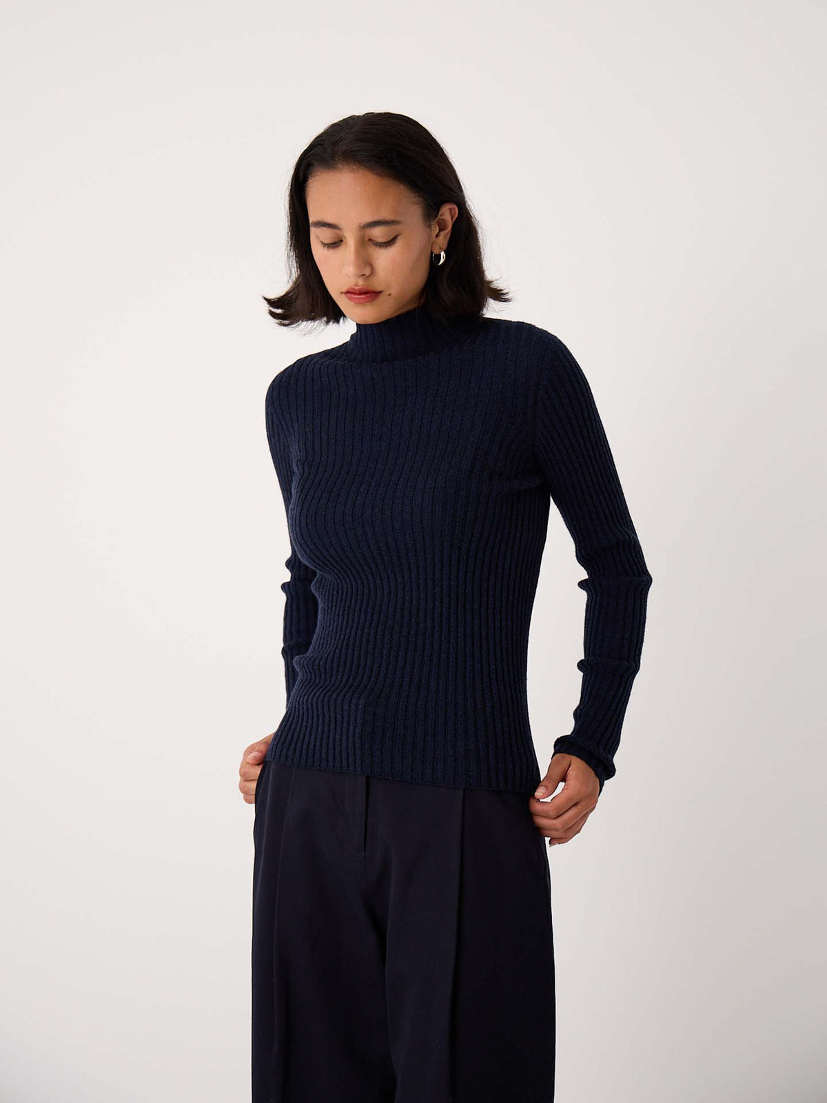 A person with shoulder-length dark hair wears the Heart Turtle Neck – Ink and dark pants, standing against a plain light background.