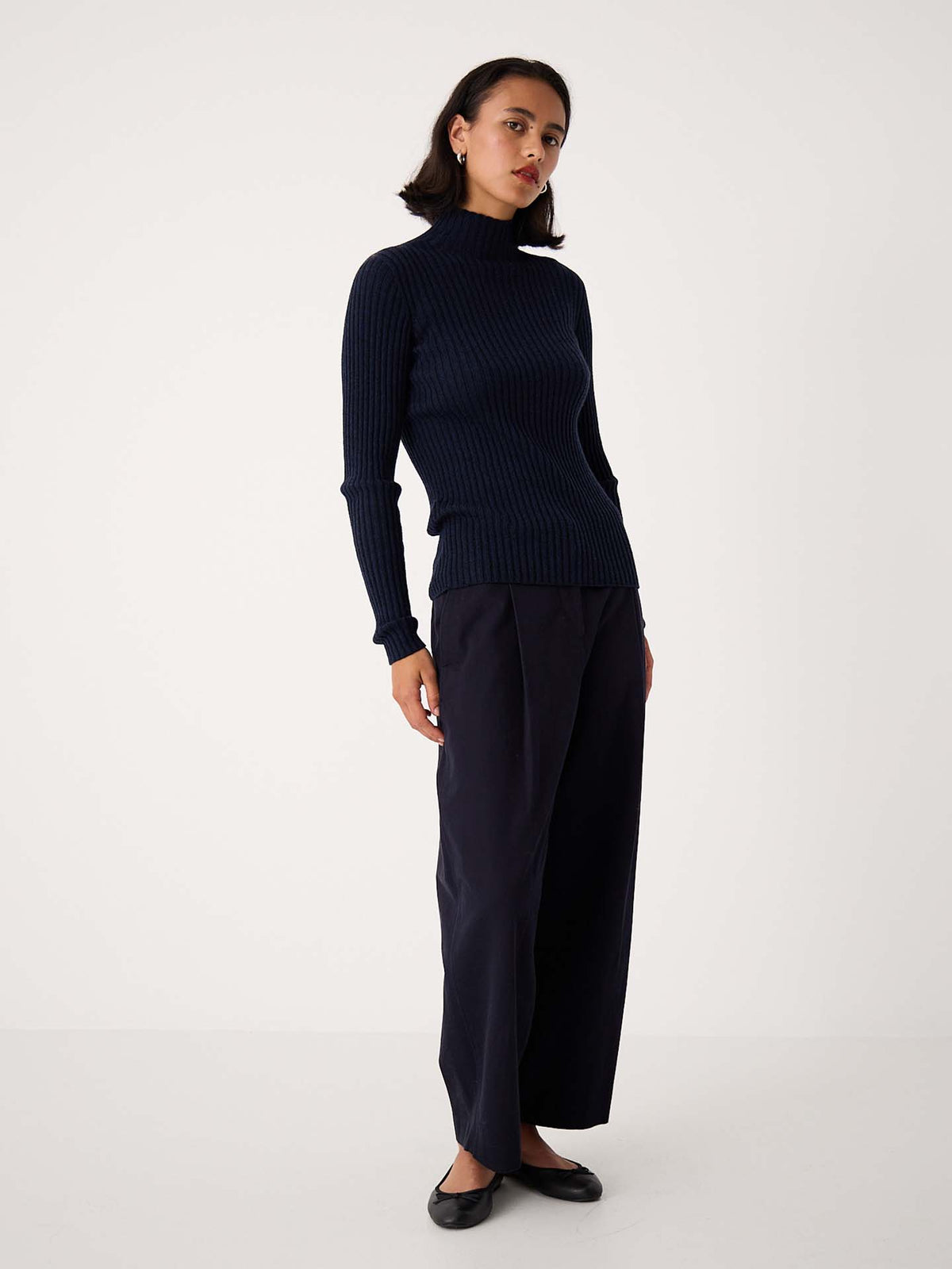 A woman stands against a plain background, wearing the Heart Turtle Neck – Ink, paired with relaxed fit wide-leg trousers and black flats.