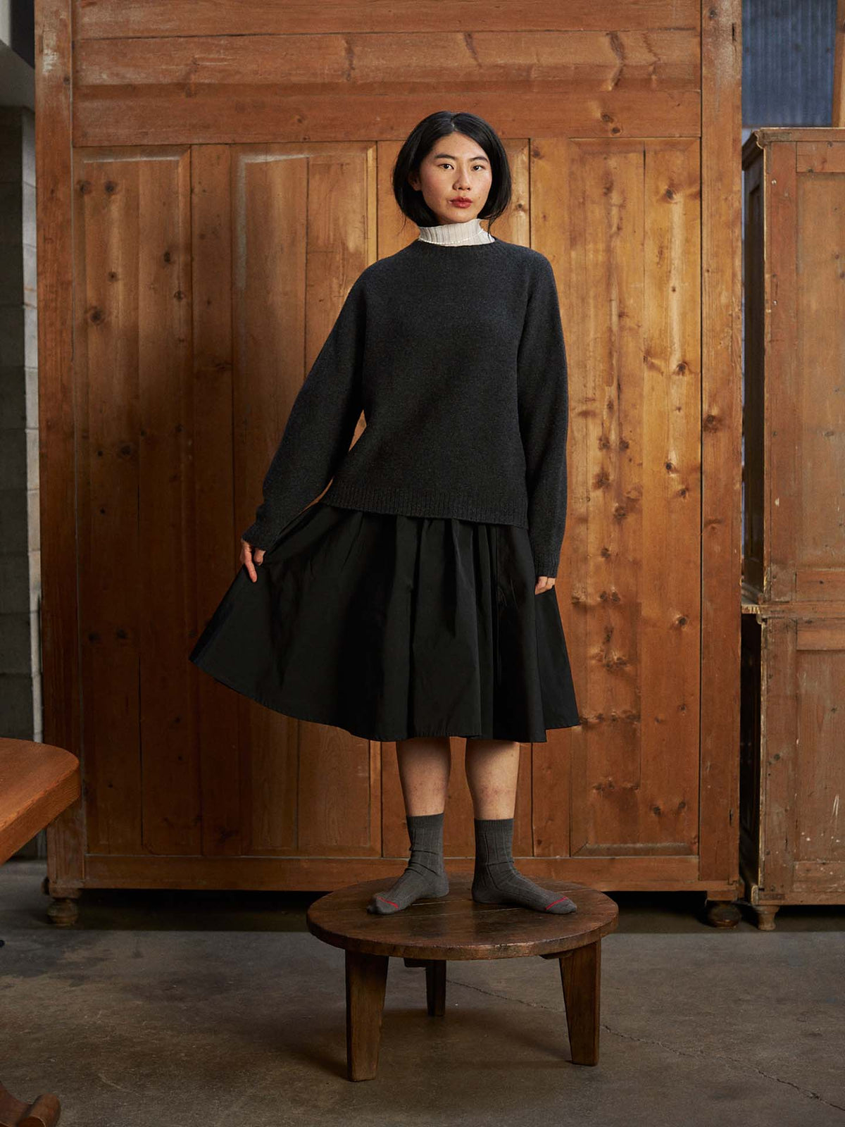 A woman stands on a round wooden stool by a wooden wall, wearing the Heart Turtle Neck – Ivory under a relaxed fit dark sweater, paired with a black skirt and gray socks.