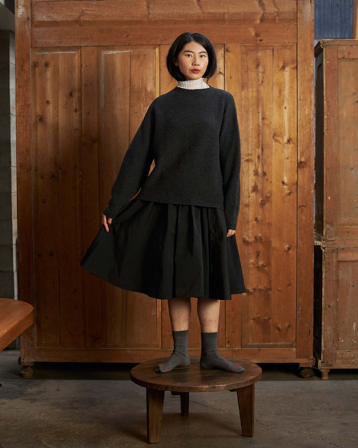 A woman stands on a round wooden stool by a wooden wall, wearing the Heart Turtle Neck – Ivory under a relaxed fit dark sweater, paired with a black skirt and gray socks.