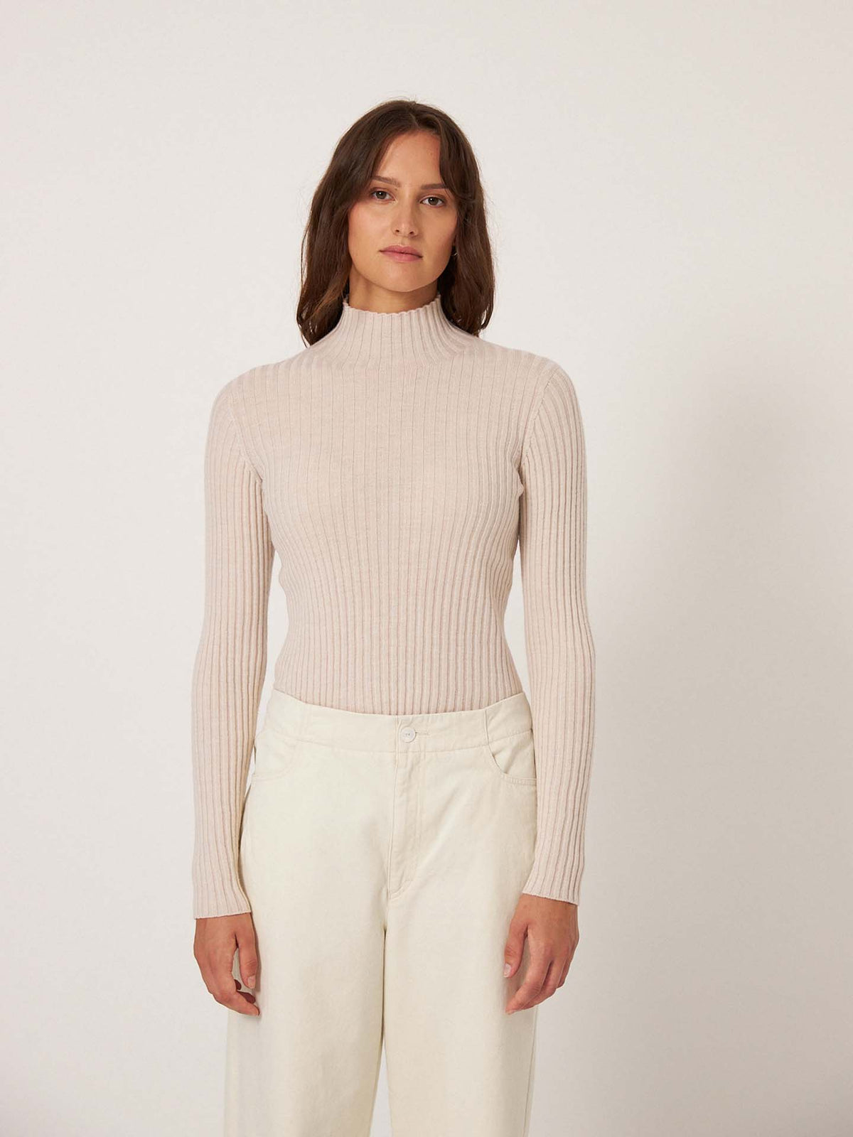 A woman faces forward against a plain background, wearing the Heart Turtle Neck – Ivory, a relaxed fit ribbed turtleneck sweater, paired with off-white pants.