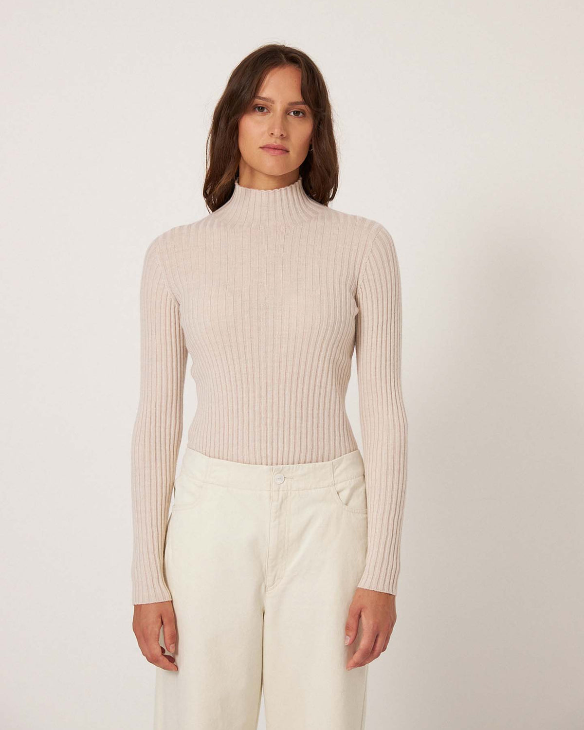 A woman faces forward against a plain background, wearing the Heart Turtle Neck – Ivory, a relaxed fit ribbed turtleneck sweater, paired with off-white pants.
