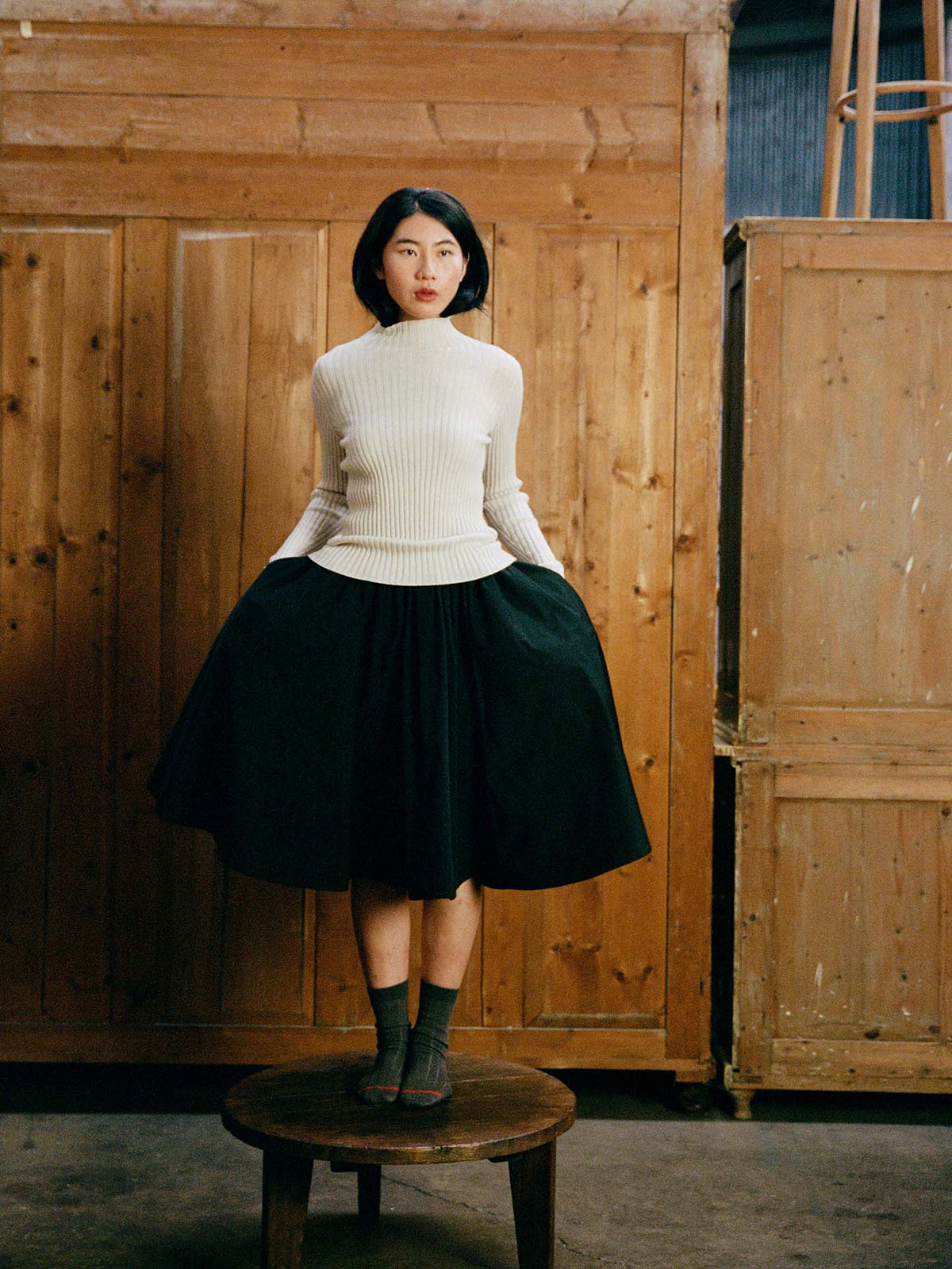A woman stands on a wooden stool against a wooden backdrop, wearing the Heart Turtle Neck – Ivory, a black skirt, and gray socks, with her hands in her pockets.