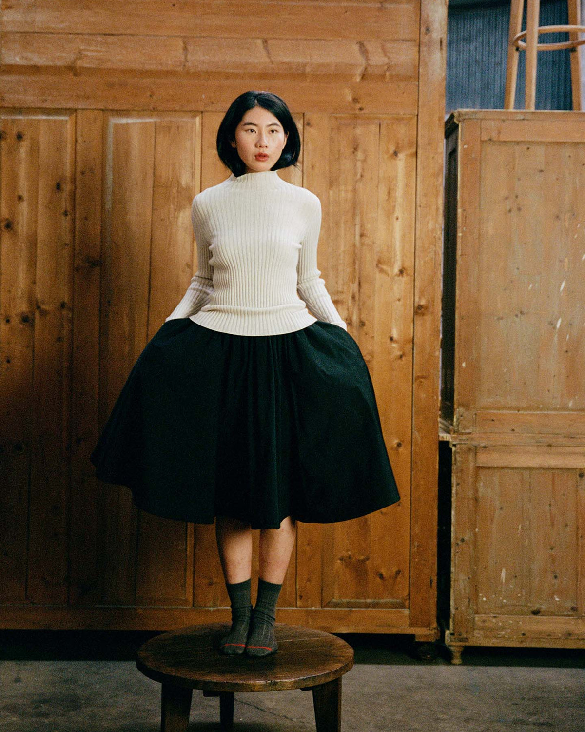 A woman stands on a wooden stool against a wooden backdrop, wearing the Heart Turtle Neck – Ivory, a black skirt, and gray socks, with her hands in her pockets.