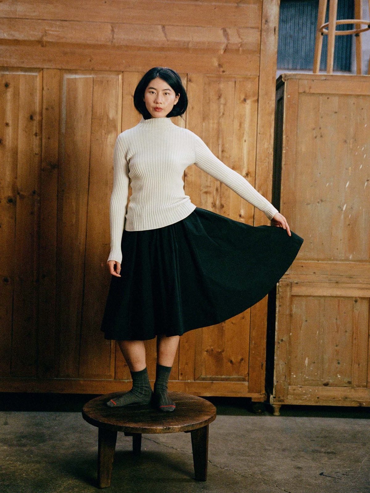 A woman wears the Heart Turtle Neck – Ivory with a black skirt, standing on a round wooden stool indoors and holding out one side of her skirt. Wooden panels are in the background.