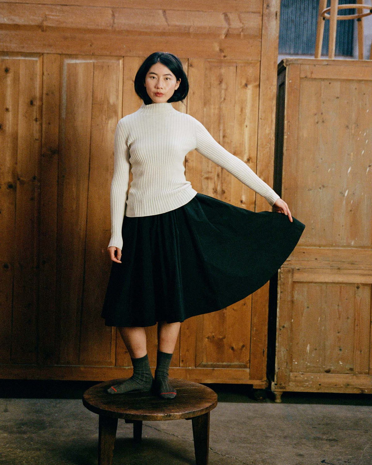 A woman wears the Heart Turtle Neck – Ivory with a black skirt, standing on a round wooden stool indoors and holding out one side of her skirt. Wooden panels are in the background.