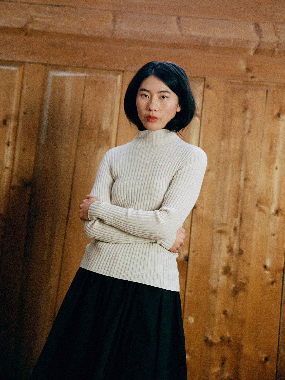 A woman with black hair, arms crossed, wears the Heart Turtle Neck – Ivory and a black skirt while standing in front of a wooden wall.