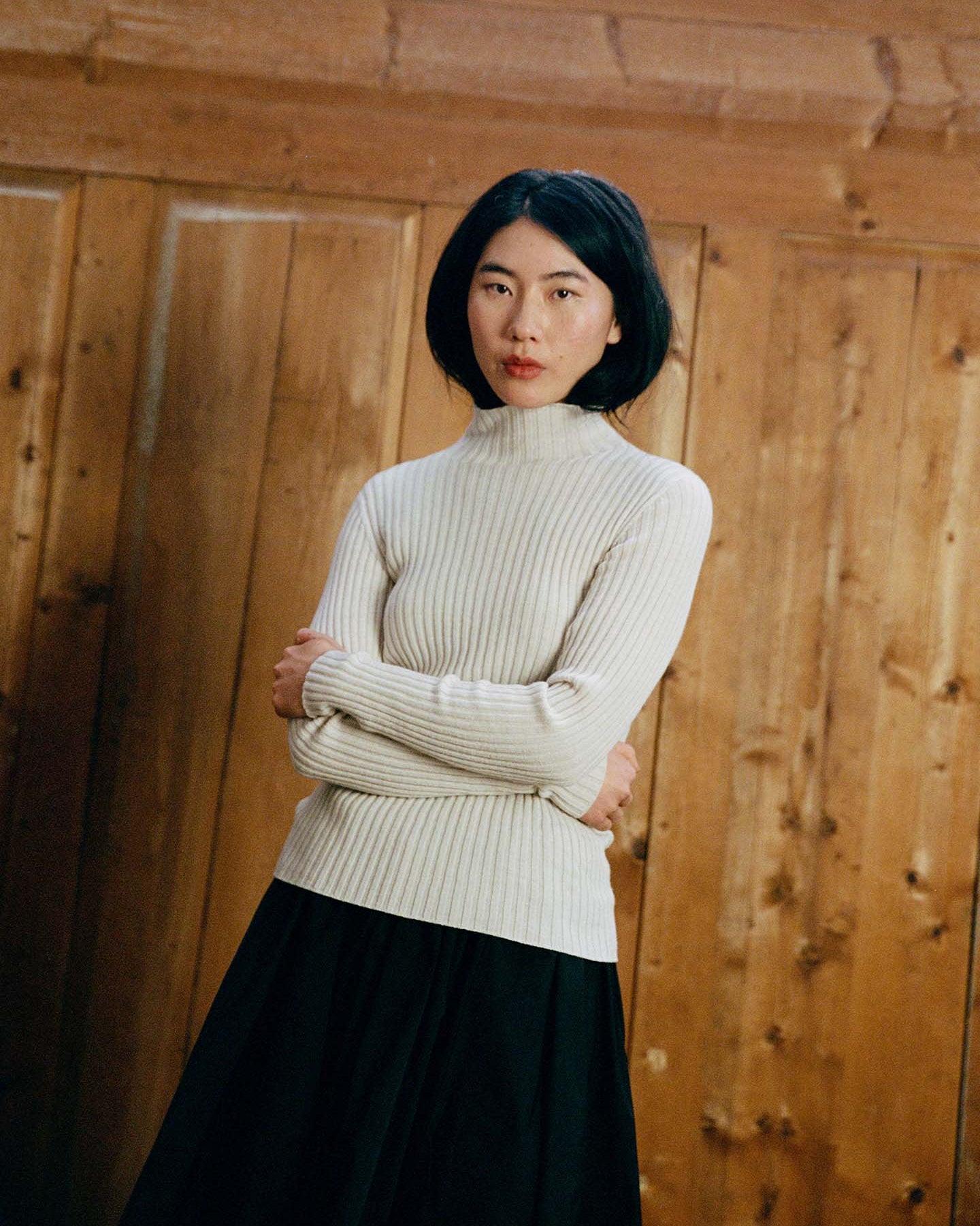 A woman with black hair, arms crossed, wears the Heart Turtle Neck – Ivory and a black skirt while standing in front of a wooden wall.