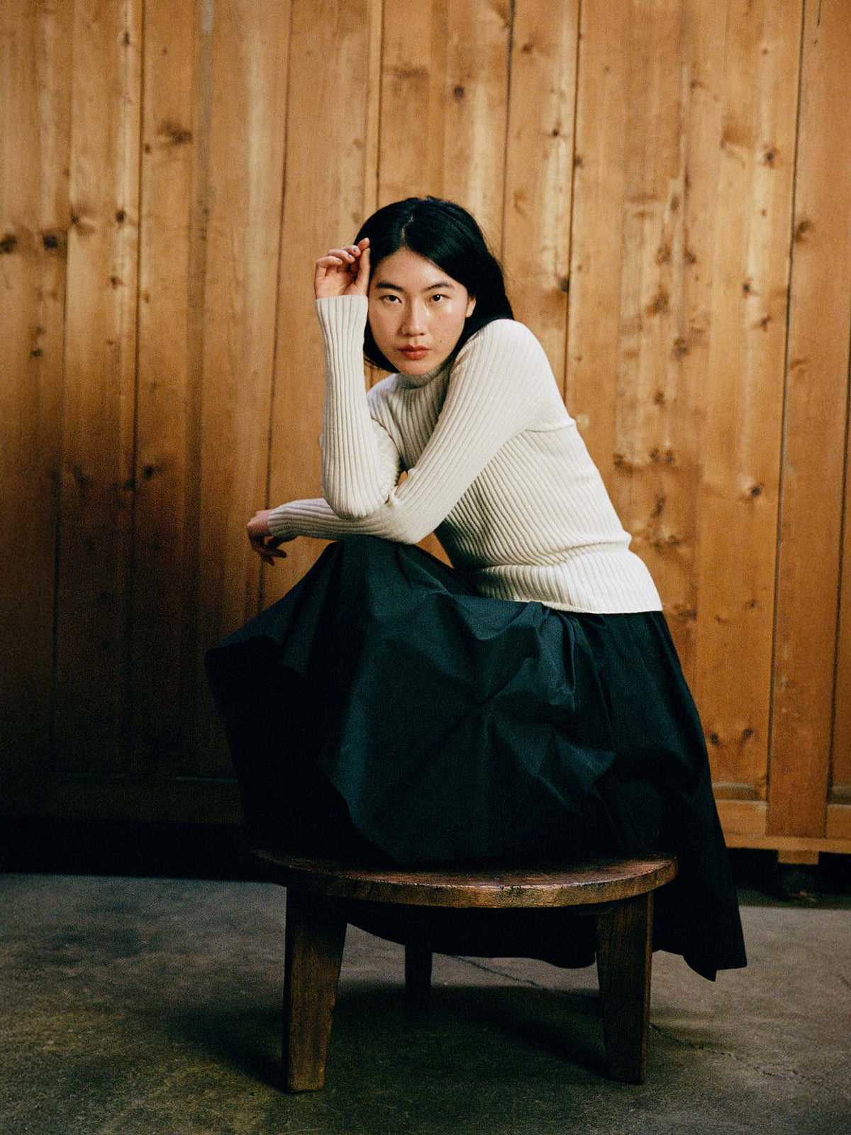 A person wearing the Heart Turtle Neck – Ivory and a long black skirt sits on a round wooden stool before a wooden wall, resting their head on one hand and looking at the camera.