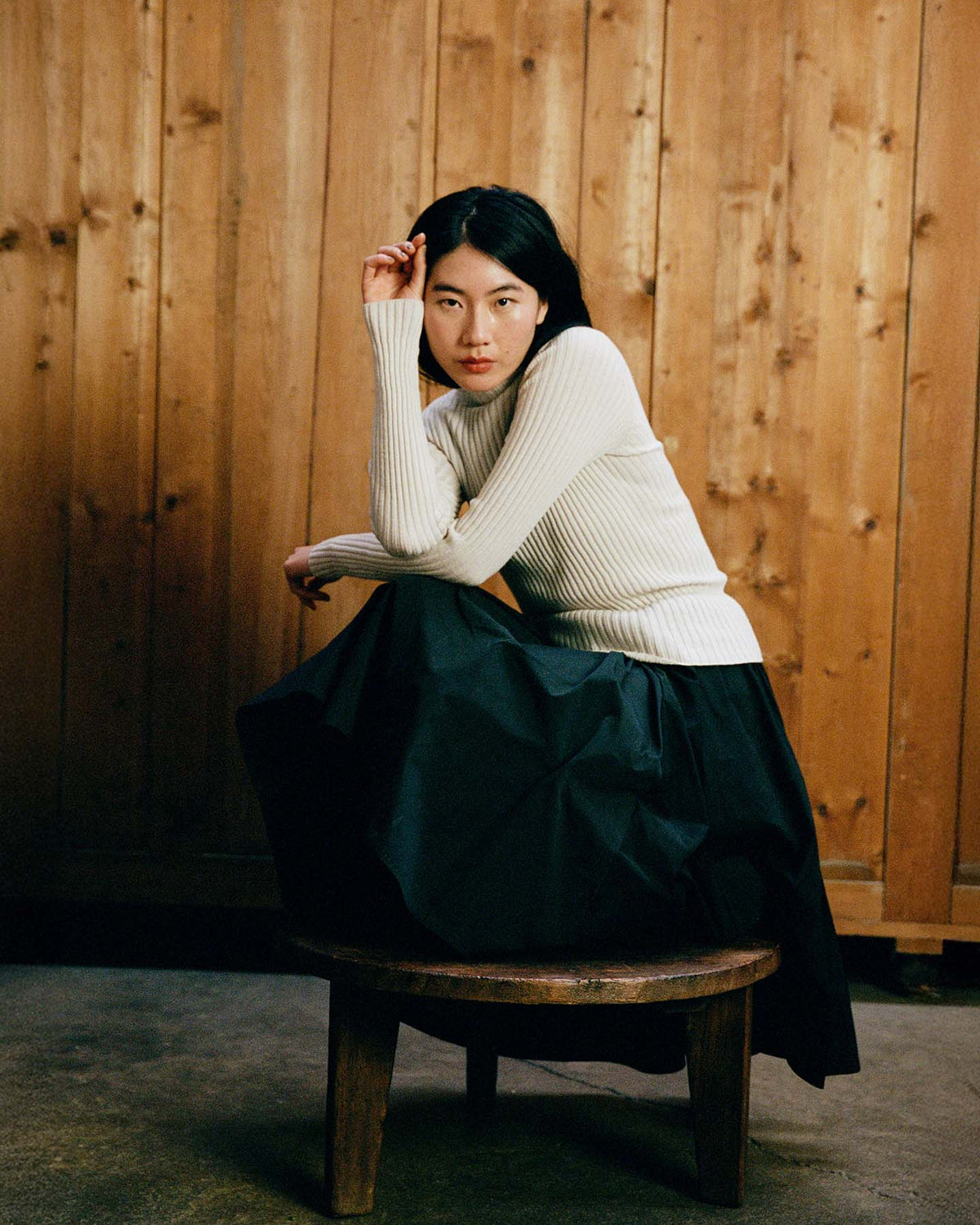 A person wearing the Heart Turtle Neck – Ivory and a long black skirt sits on a round wooden stool before a wooden wall, resting their head on one hand and looking at the camera.