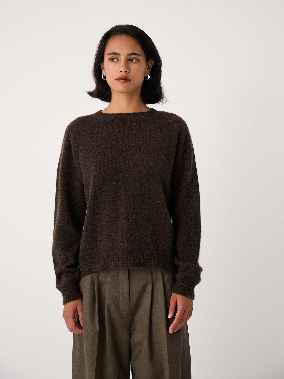 A woman stands against a plain background, wearing the Nimbus Raglan Knit in Truffle with relaxed fit, high-waisted olive green trousers.