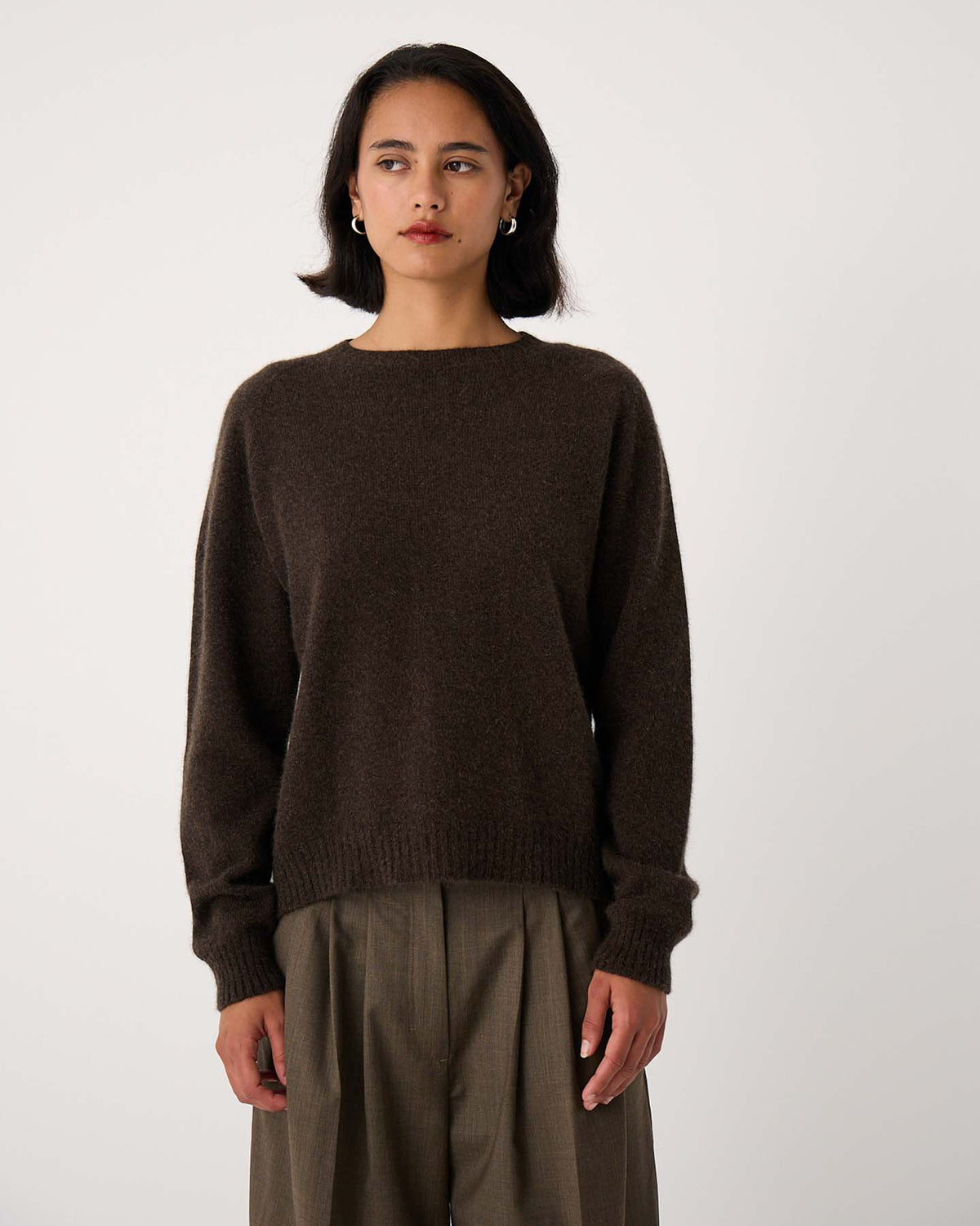 A woman stands against a plain background, wearing the Nimbus Raglan Knit in Truffle with relaxed fit, high-waisted olive green trousers.