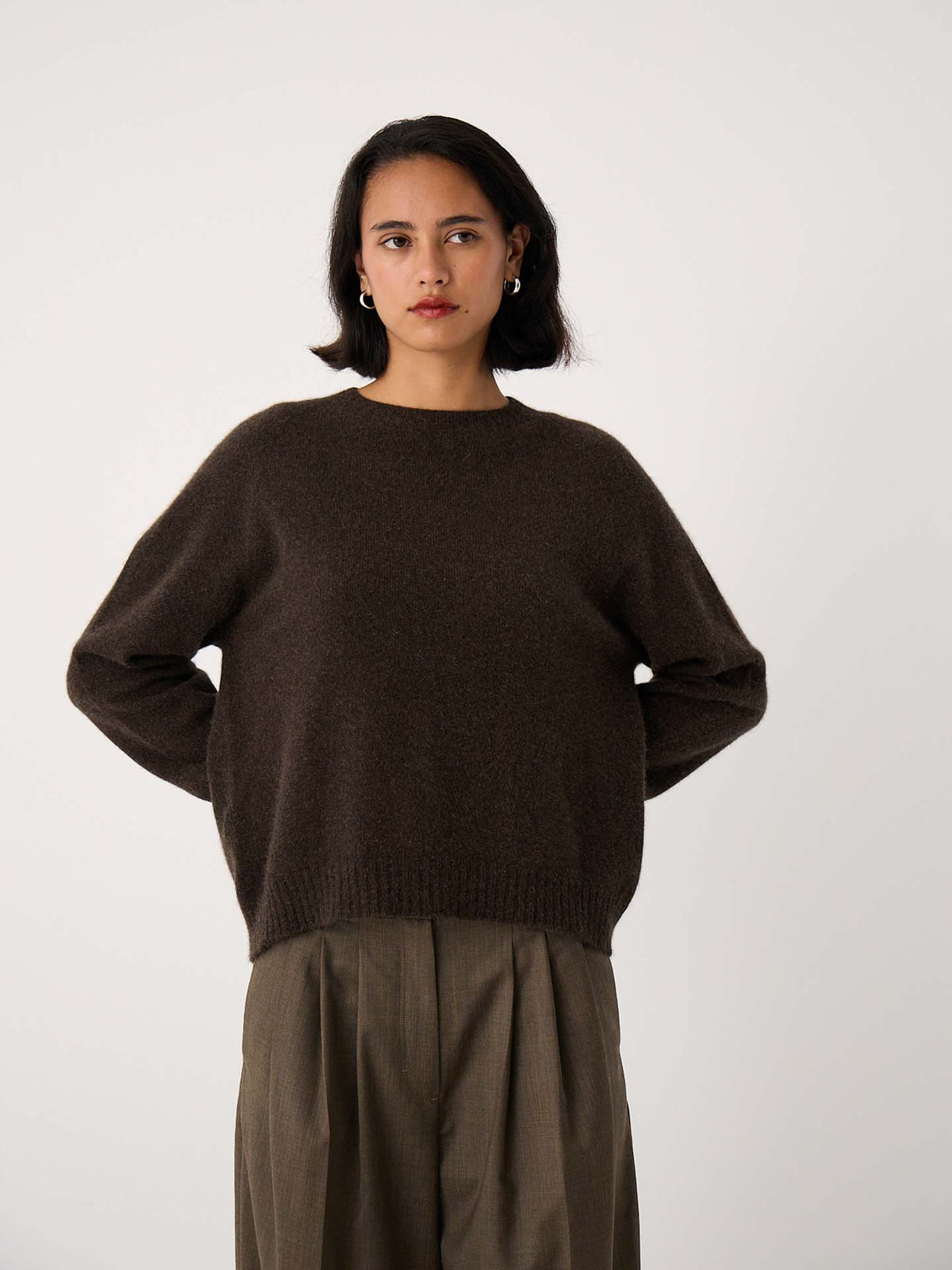 Woman stands against a plain background, wearing the Nimbus Raglan Knit in Truffle—a slightly fitted dark brown sweater—paired with loose olive trousers. Her hands are behind her back and she has a neutral expression.