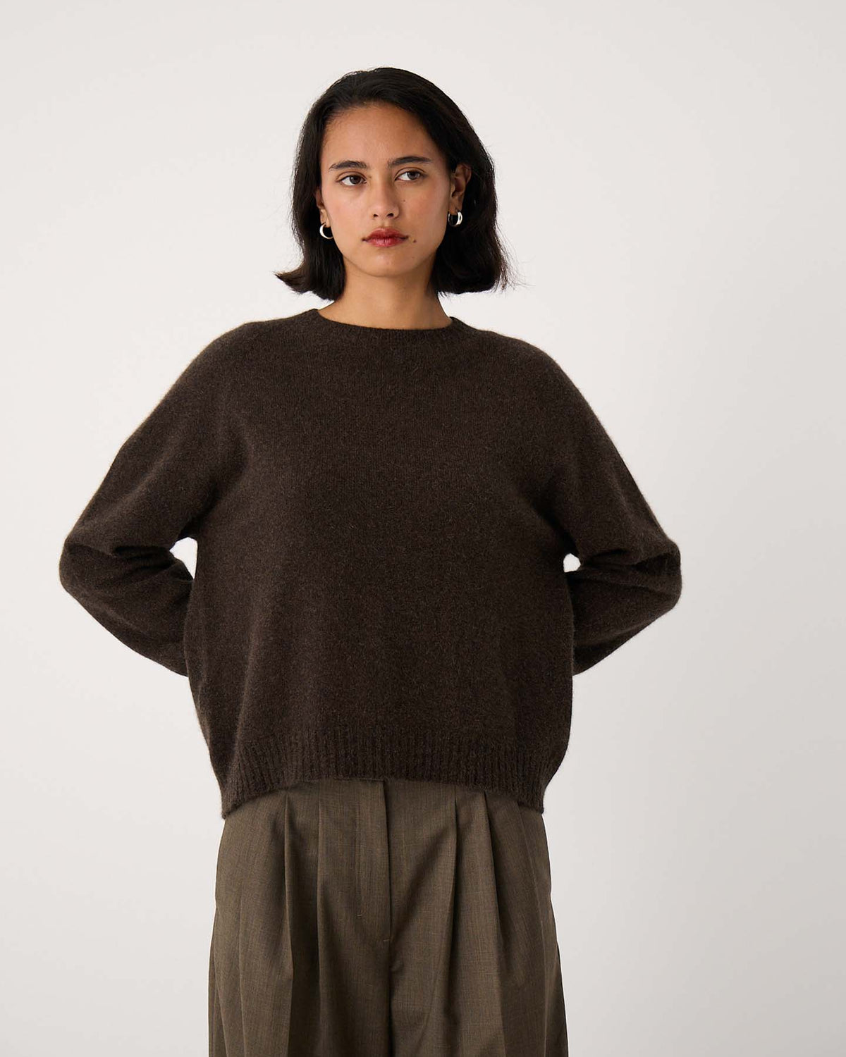 Woman stands against a plain background, wearing the Nimbus Raglan Knit in Truffle—a slightly fitted dark brown sweater—paired with loose olive trousers. Her hands are behind her back and she has a neutral expression.