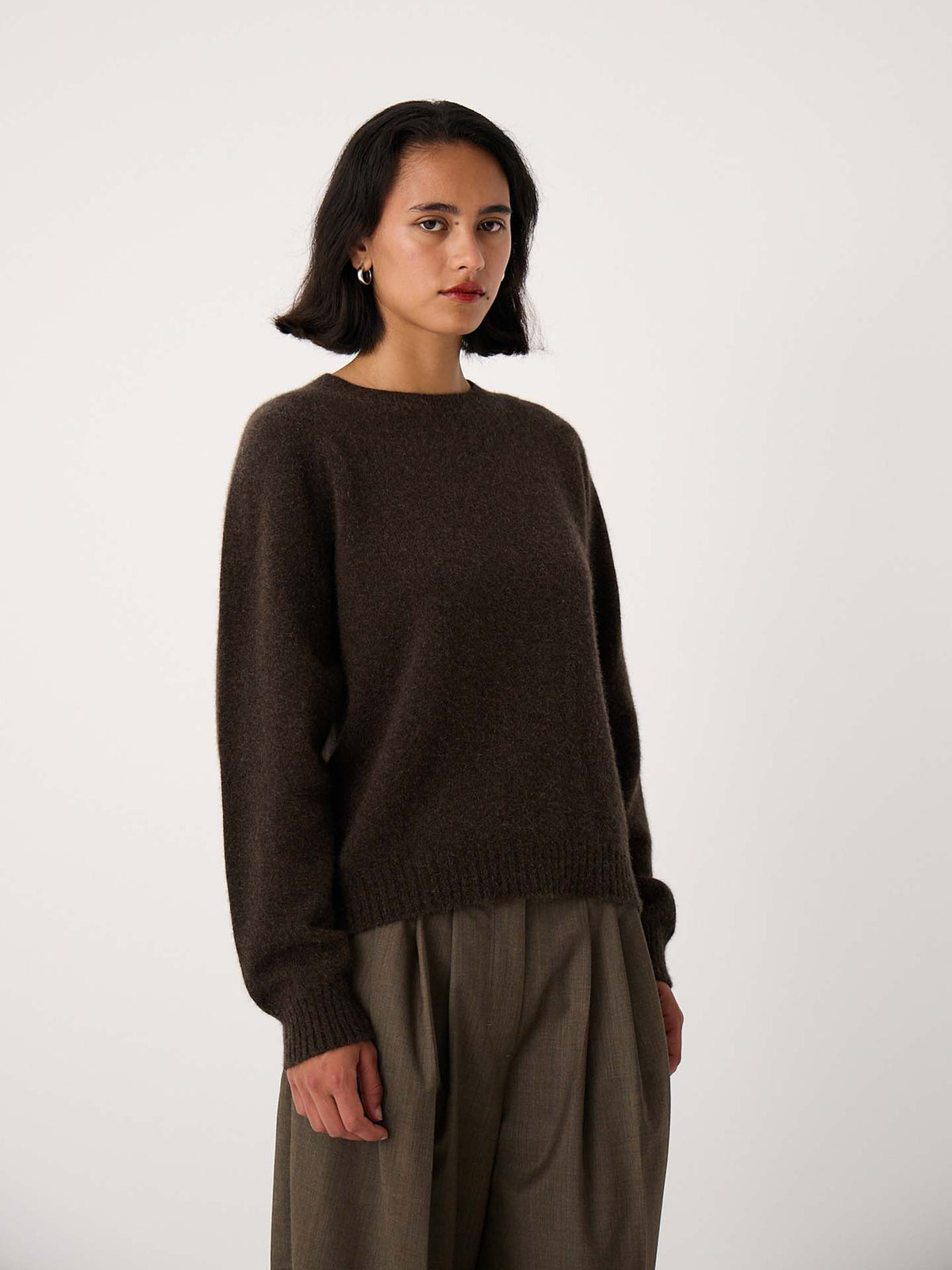 A woman with shoulder-length dark hair wears the Nimbus Raglan Knit in Truffle, a slightly fitted dark brown sweater, paired with relaxed fit olive trousers, standing against a plain light background.