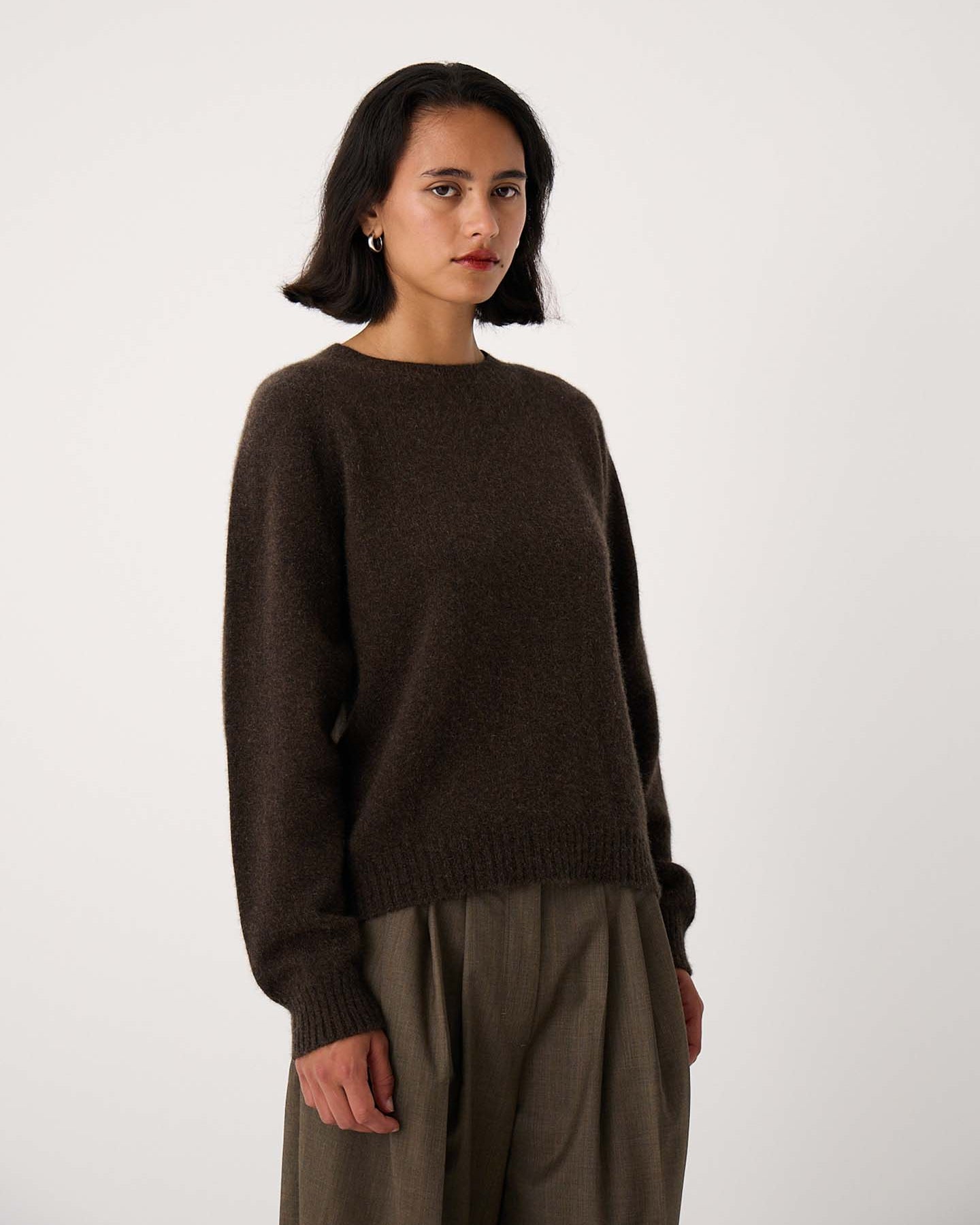 A woman with shoulder-length dark hair wears the Nimbus Raglan Knit in Truffle, a slightly fitted dark brown sweater, paired with relaxed fit olive trousers, standing against a plain light background.