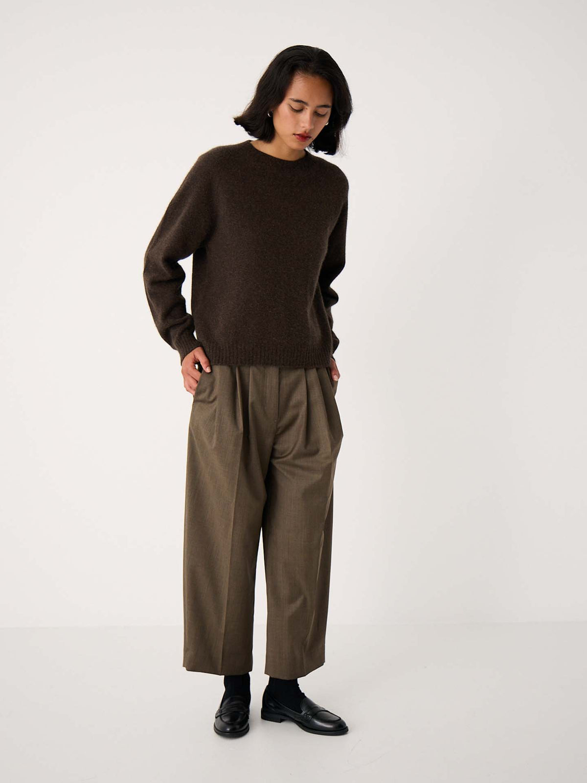 A person stands against a plain white background wearing the Nimbus Raglan Knit – Truffle sweater with relaxed-fit, wide-legged khaki trousers and black shoes, looking down with hands in pockets.