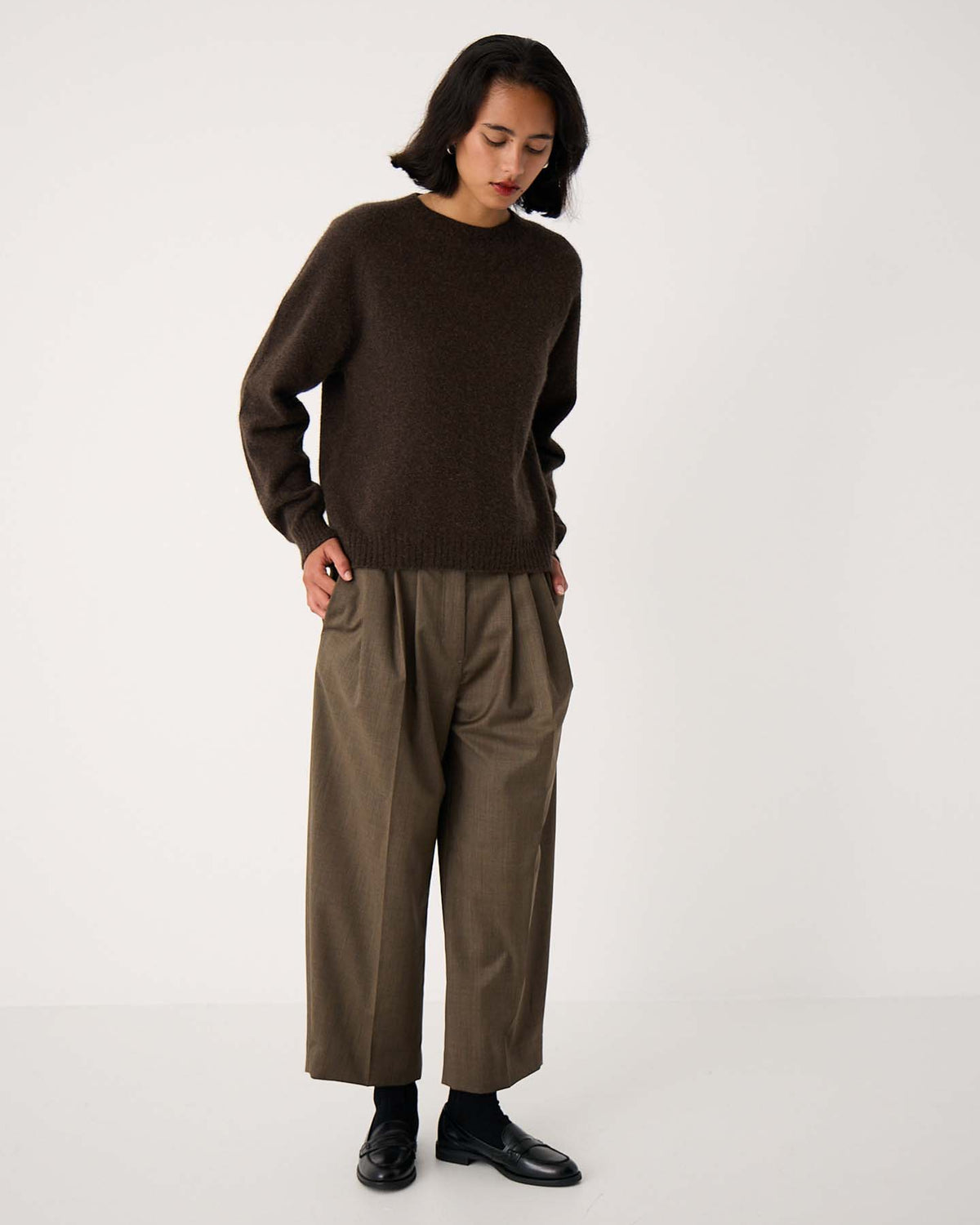 A person stands against a plain white background wearing the Nimbus Raglan Knit – Truffle sweater with relaxed-fit, wide-legged khaki trousers and black shoes, looking down with hands in pockets.