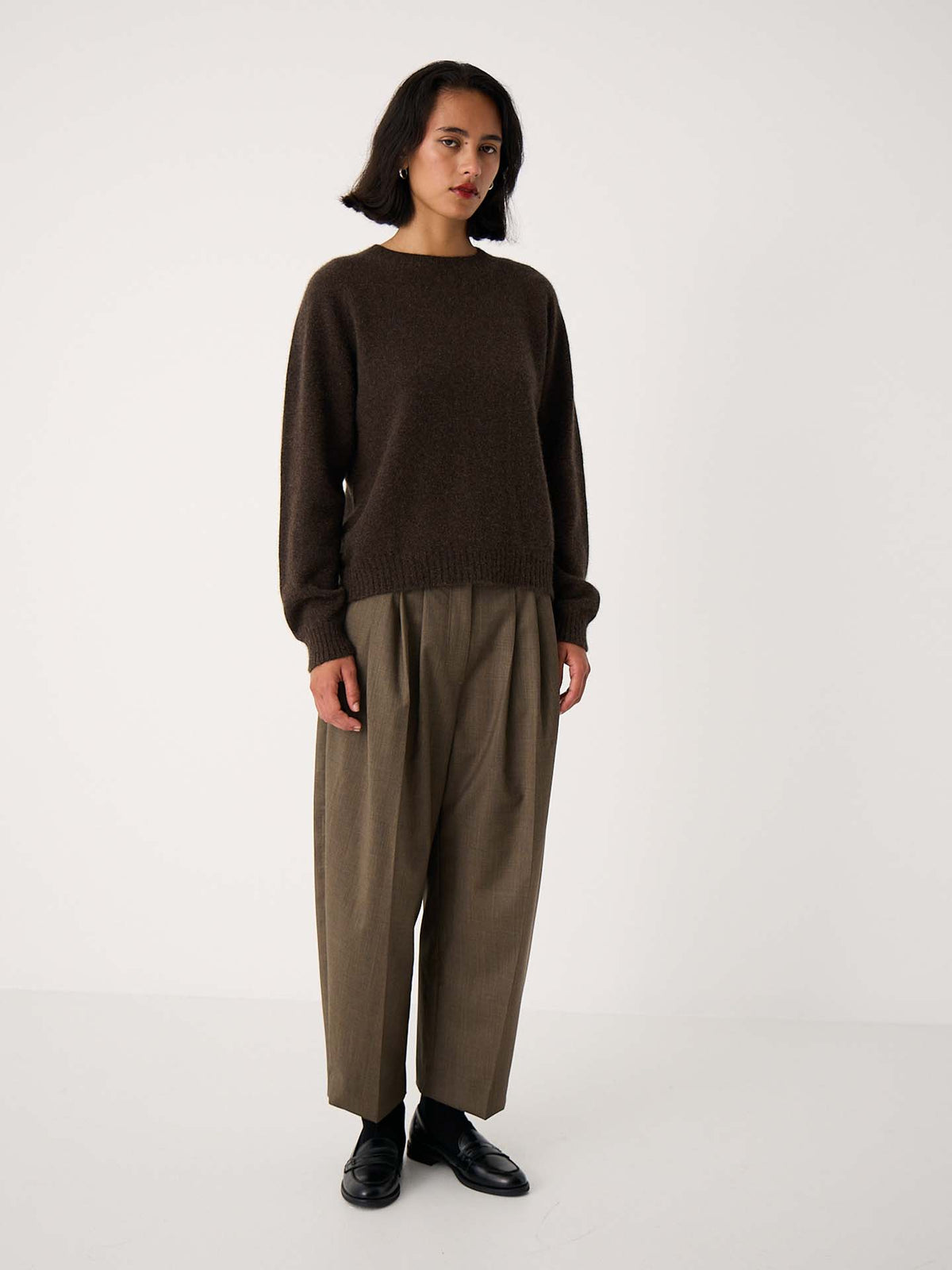 A person stands against a plain white background wearing the Nimbus Raglan Knit – Truffle, a slightly fitted dark brown sweater, paired with loose tan trousers and black shoes.