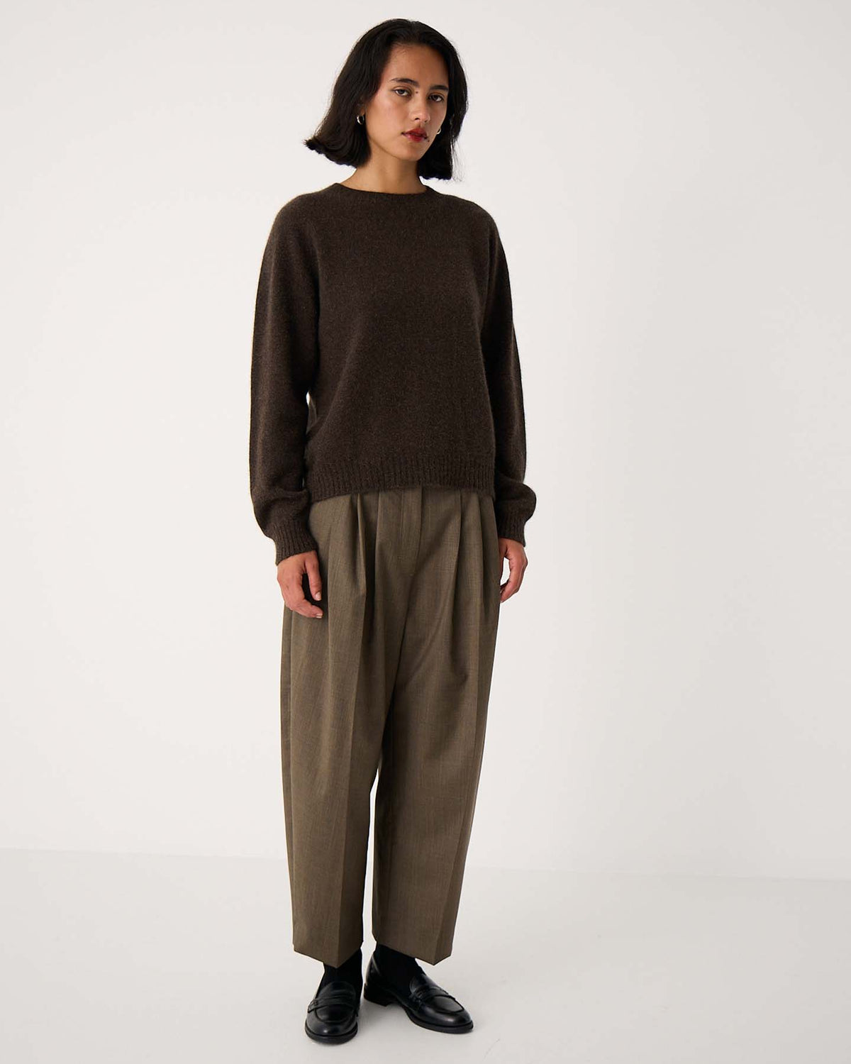 A person stands against a plain white background wearing the Nimbus Raglan Knit – Truffle, a slightly fitted dark brown sweater, paired with loose tan trousers and black shoes.