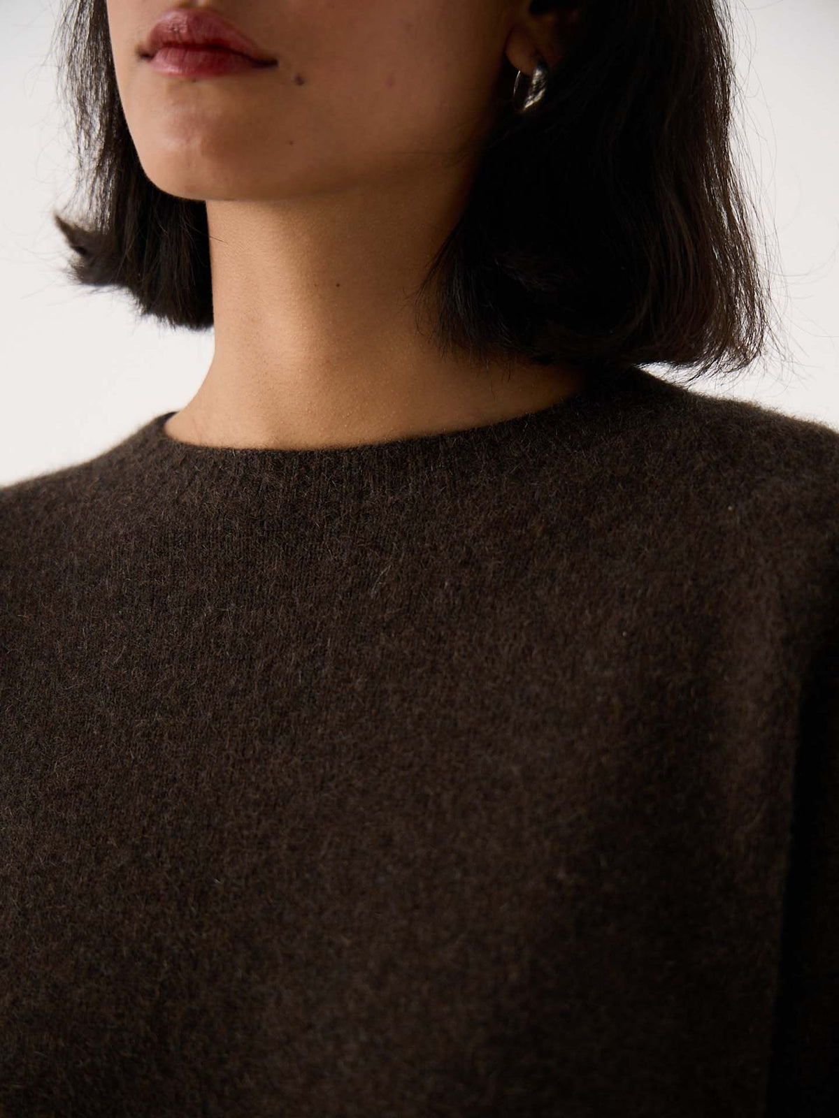 A woman with short dark hair models the Nimbus Raglan Knit – Truffle, a relaxed-fit dark brown sweater; only her lower face, neck, and upper chest are shown. She also wears a silver hoop earring.