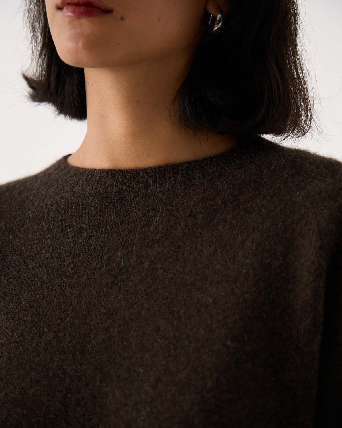 A woman with short dark hair models the Nimbus Raglan Knit – Truffle, a relaxed-fit dark brown sweater; only her lower face, neck, and upper chest are shown. She also wears a silver hoop earring.