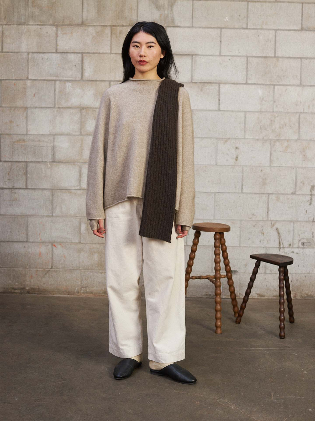 A woman stands indoors against a cinder block wall, wearing a beige sweater, white wide-leg pants, black shoes, and the Ripple Scarf – Truffle (170cm) draped over one shoulder. Two wooden stools are visible in the background.