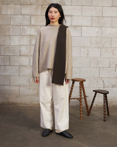 A woman stands indoors against a cinder block wall, wearing a beige sweater, white wide-leg pants, black shoes, and the Ripple Scarf – Truffle (170cm) draped over one shoulder. Two wooden stools are visible in the background.