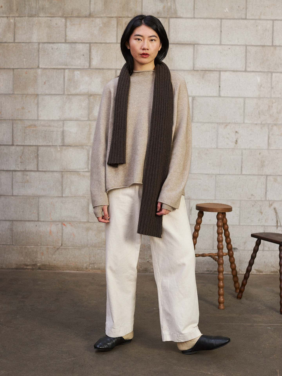 A person stands indoors by a cinder block wall, wearing a beige sweater, wide white pants, black shoes, and the Ripple Scarf – Truffle (170cm). Two wooden stools are nearby.