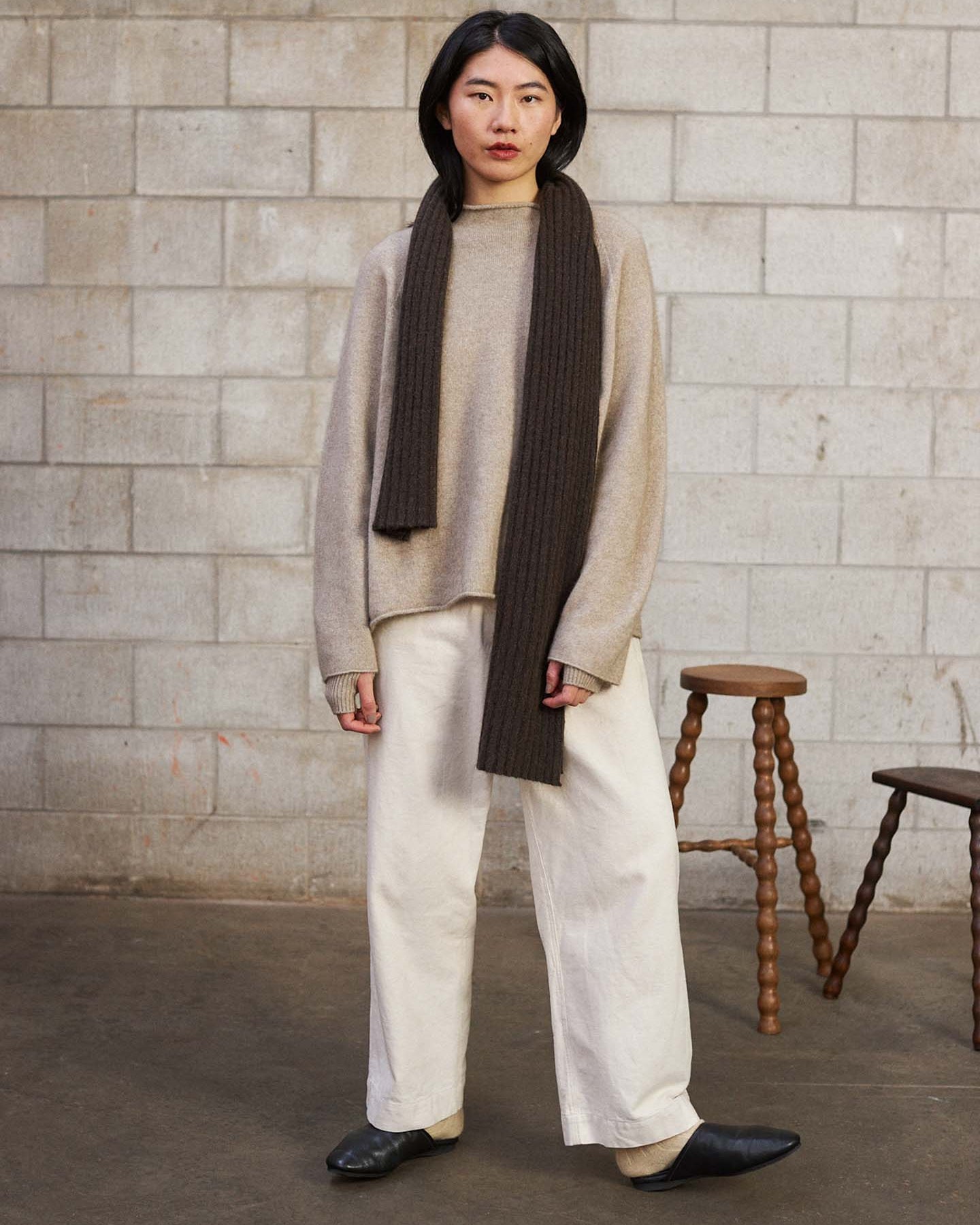 A woman stands indoors against a cinder block wall, wearing a beige sweater, white wide-leg pants, black shoes, and the Ripple Scarf – Truffle (170cm) draped over one shoulder. Two wooden stools are visible in the background.