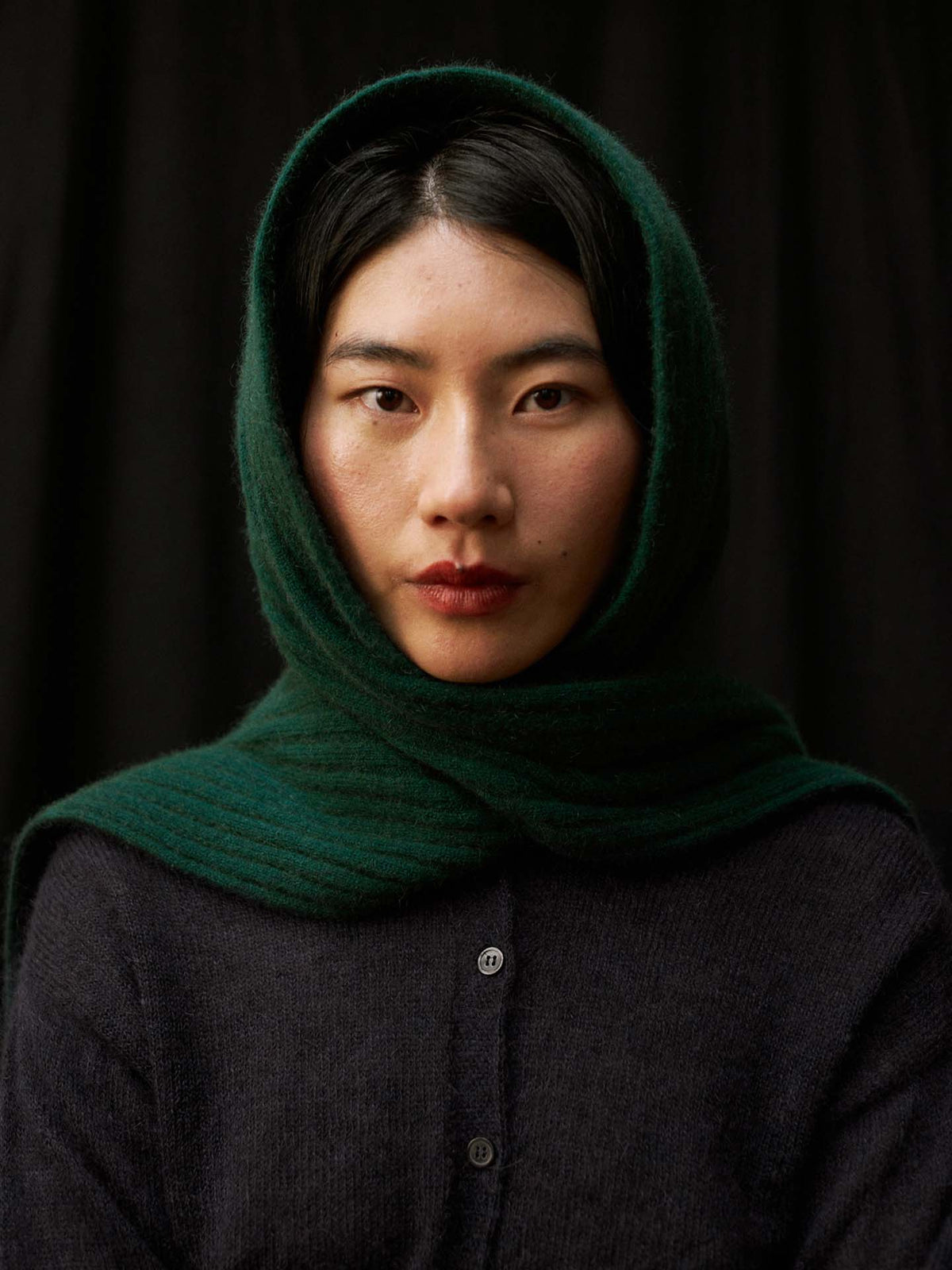 A woman with straight black hair, wearing the Ripple Scarf – Verdant Green draped over her head and a dark sweater, looks directly at the camera against a black background.