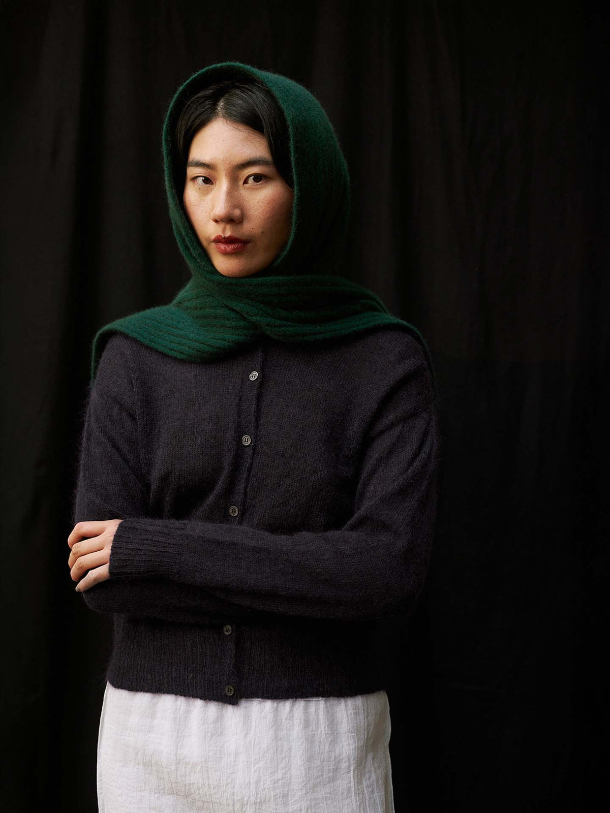 A person with long dark hair drapes the Ripple Scarf – Verdant Green over their head, pairing it with a dark button-up sweater and white skirt, standing against a plain black background.