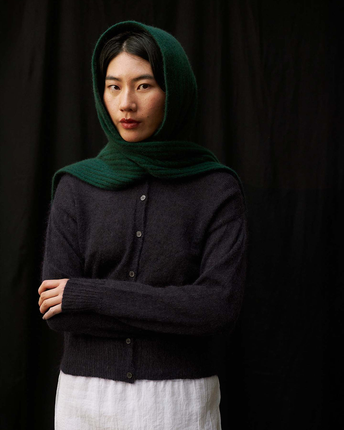 A person with long dark hair drapes the Ripple Scarf – Verdant Green over their head, pairing it with a dark button-up sweater and white skirt, standing against a plain black background.