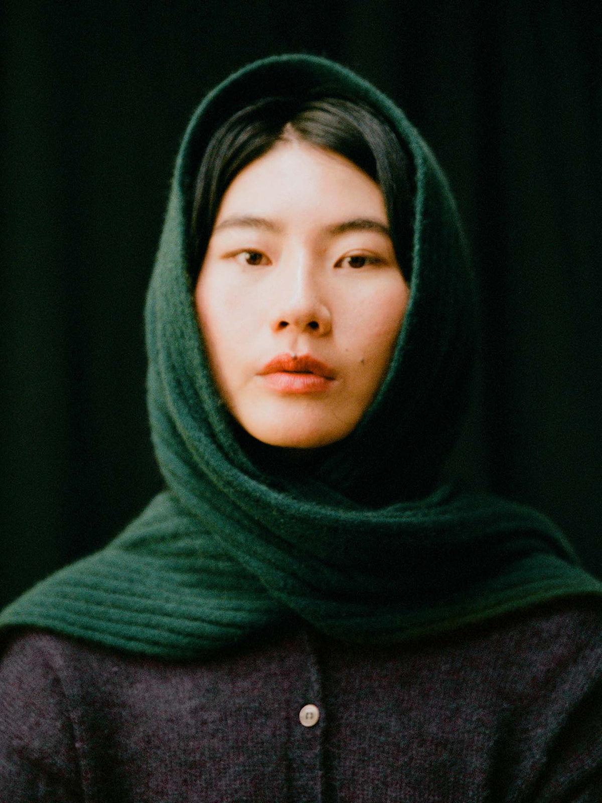 A person with straight black hair wears the 170cm Ripple Scarf – Verdant Green over their head and shoulders, paired with a dark top, set against a black background.