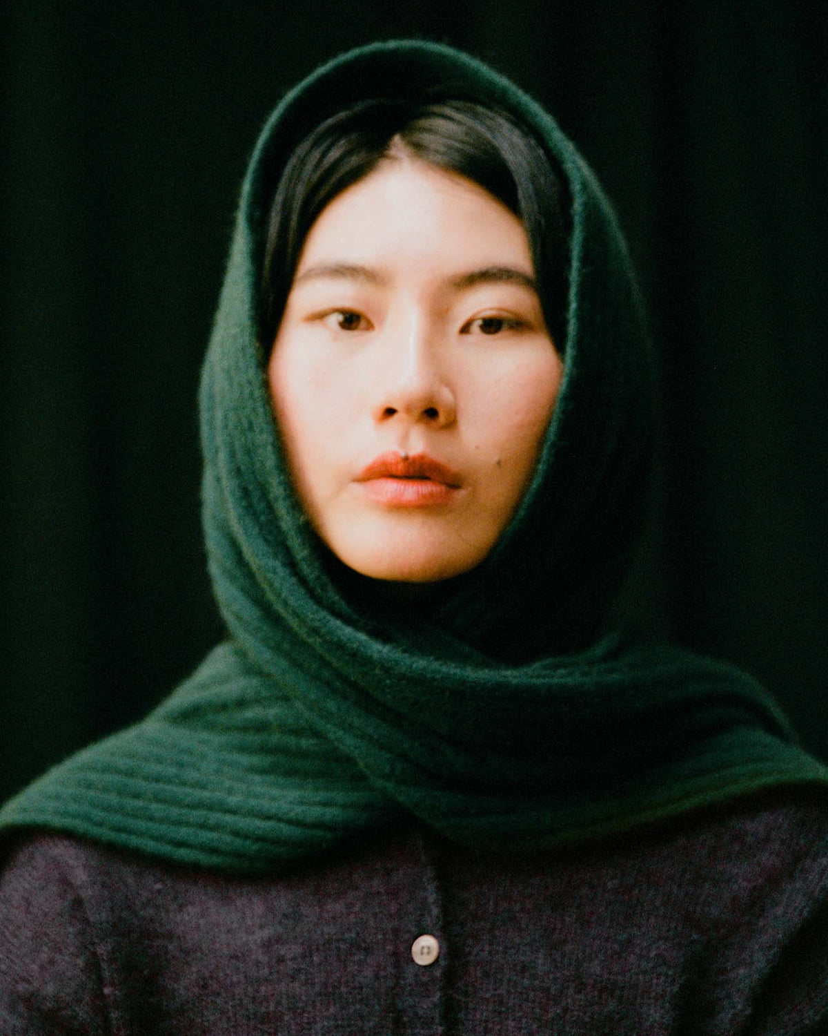 A person with straight black hair wears the 170cm Ripple Scarf – Verdant Green over their head and shoulders, paired with a dark top, set against a black background.