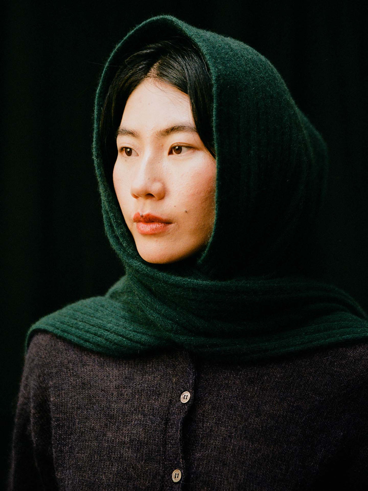 A person with straight dark hair styles the Ripple Scarf – Verdant Green, paired with a dark green hooded shawl and buttoned dark sweater, looking slightly to the side against a black background.