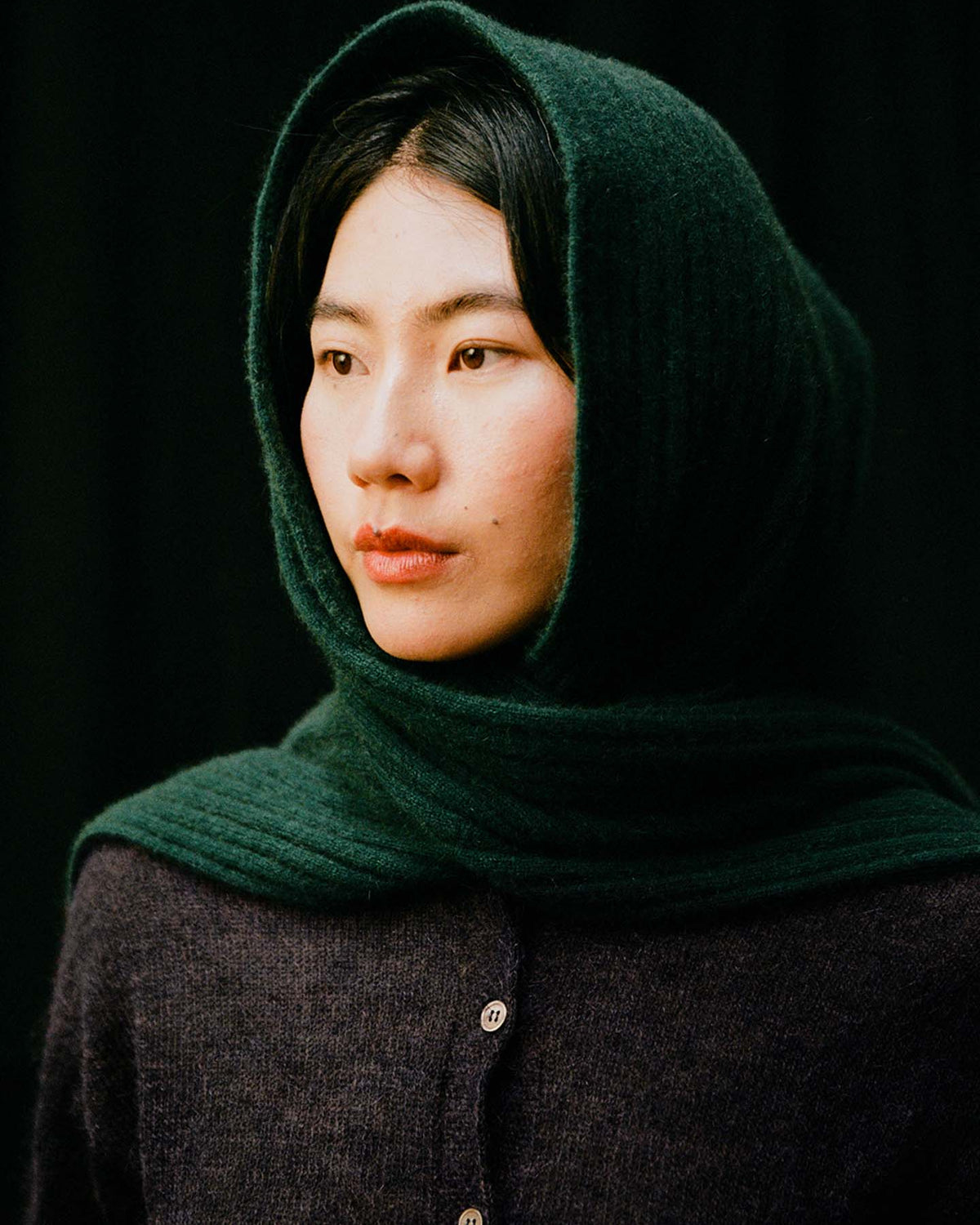 A person with straight dark hair styles the Ripple Scarf – Verdant Green, paired with a dark green hooded shawl and buttoned dark sweater, looking slightly to the side against a black background.