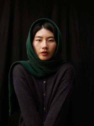 A person wears the Ripple Scarf – Verdant Green as a headscarf with a dark buttoned sweater, posing against a black background and looking directly at the camera.
