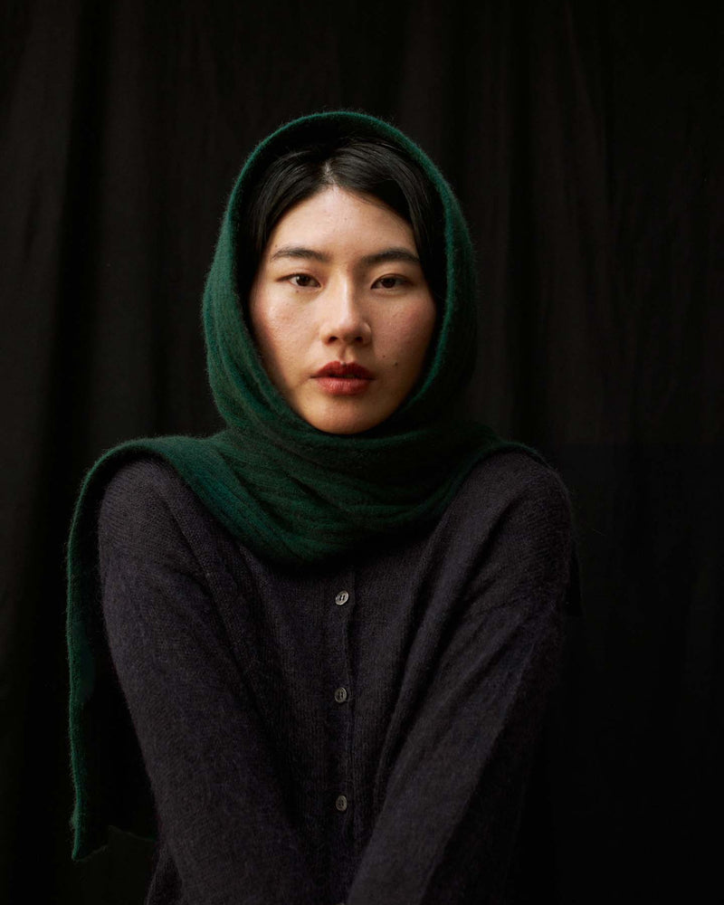 A person wears the Ripple Scarf – Verdant Green as a headscarf with a dark buttoned sweater, posing against a black background and looking directly at the camera.